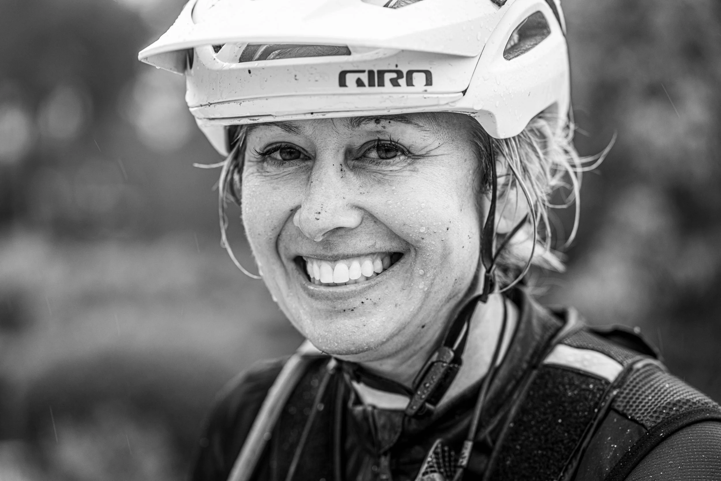 Close-up of a smiling woman wearing a bike helmet, with water droplets on her face and wet hair, outdoors.