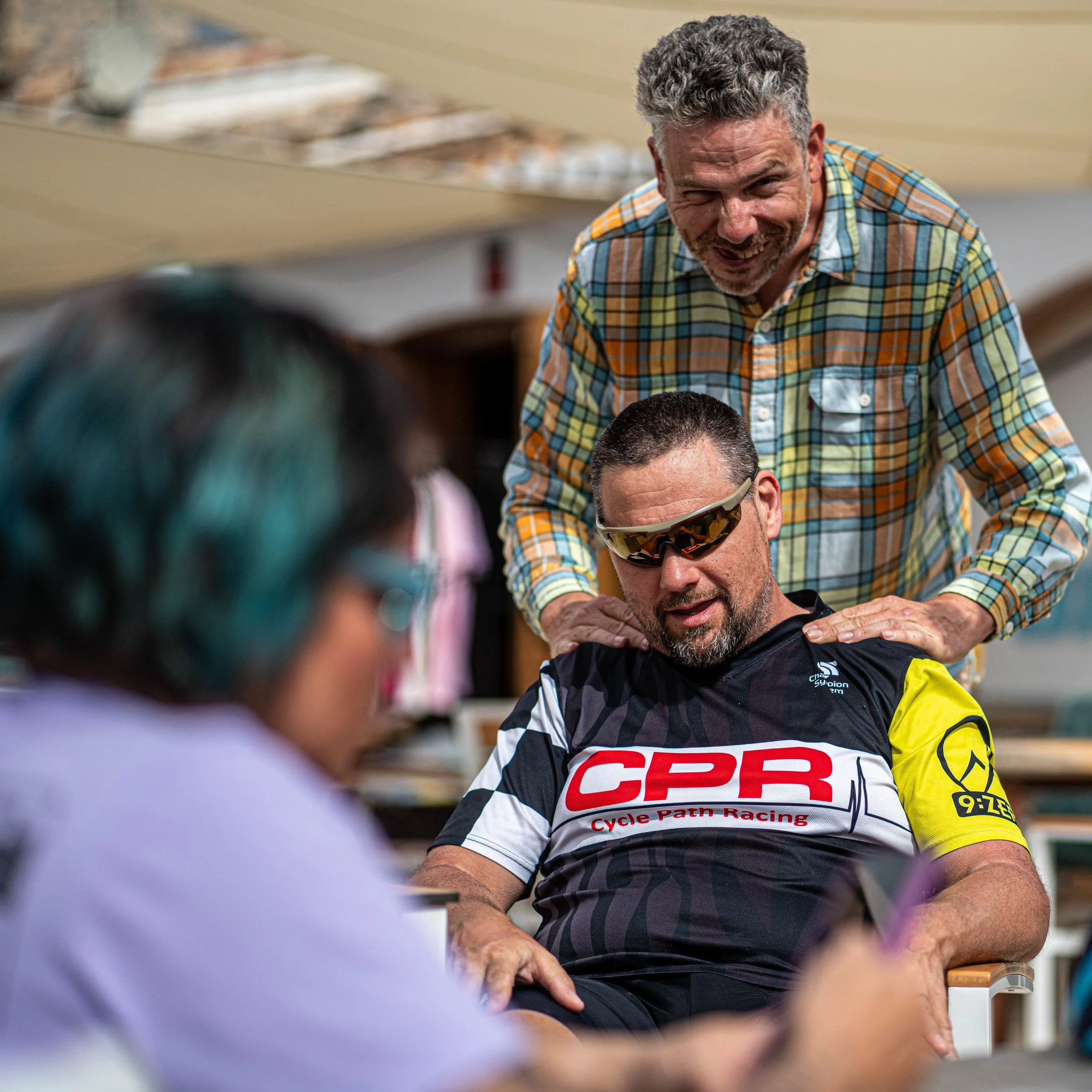 A man wearing sunglasses and a cycling jersey receiving a shoulder massage from another man in a plaid shirt, with a woman in the foreground looking at a phone.