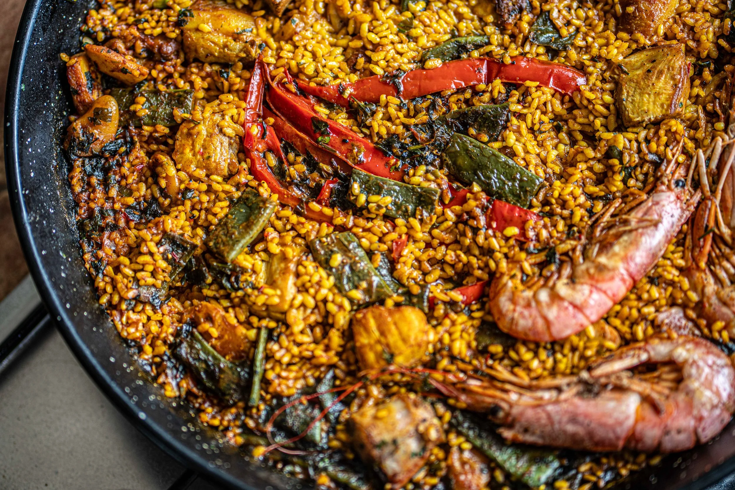 A close-up of a skillet filled with cooked seafood paella, featuring rice, shrimp, red peppers, shellfish, and green vegetables.