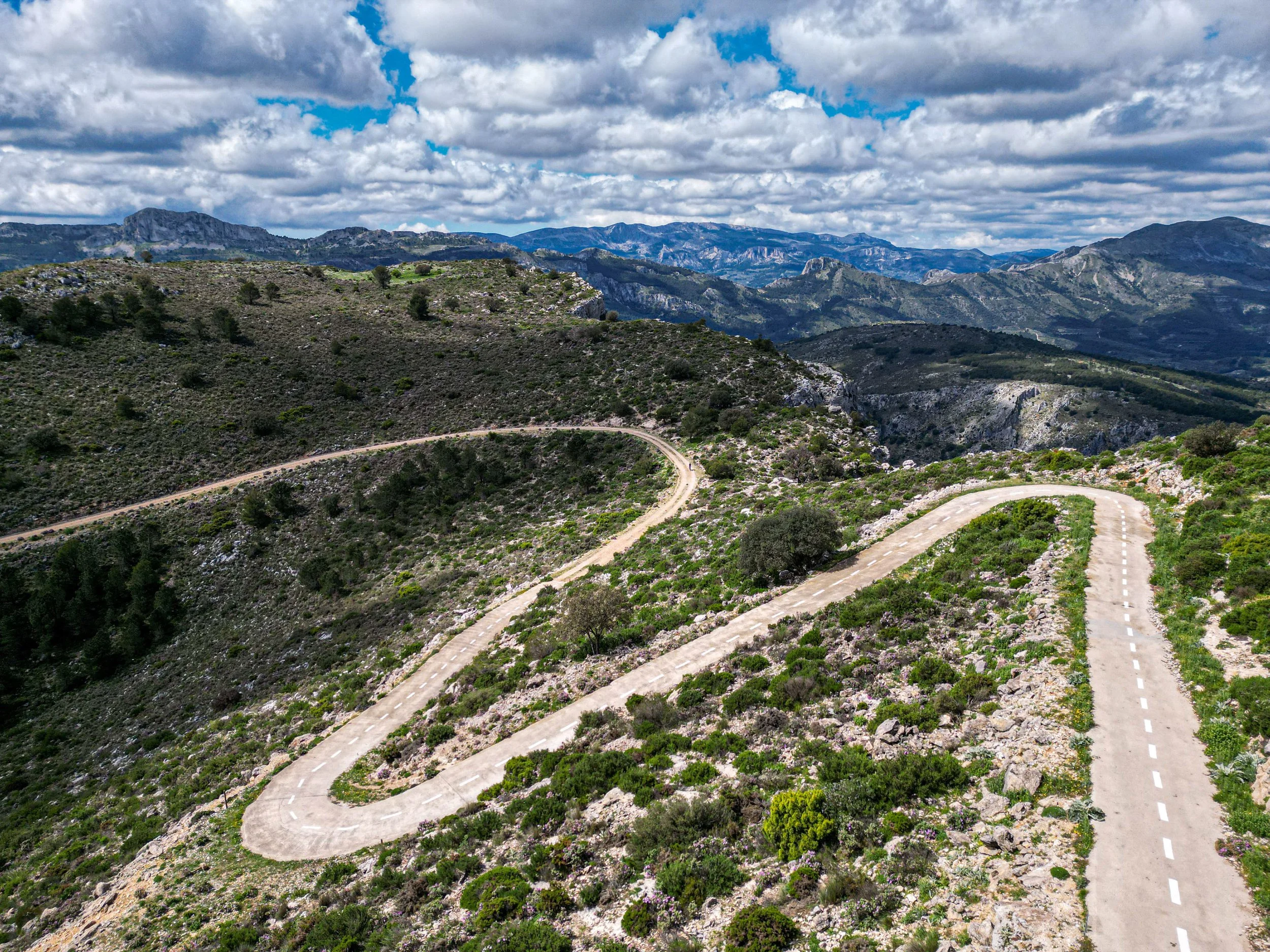 A winding mountain road with white dashed lines through rugged terrain with green shrubs and rocky outcrops, under a partly cloudy sky.