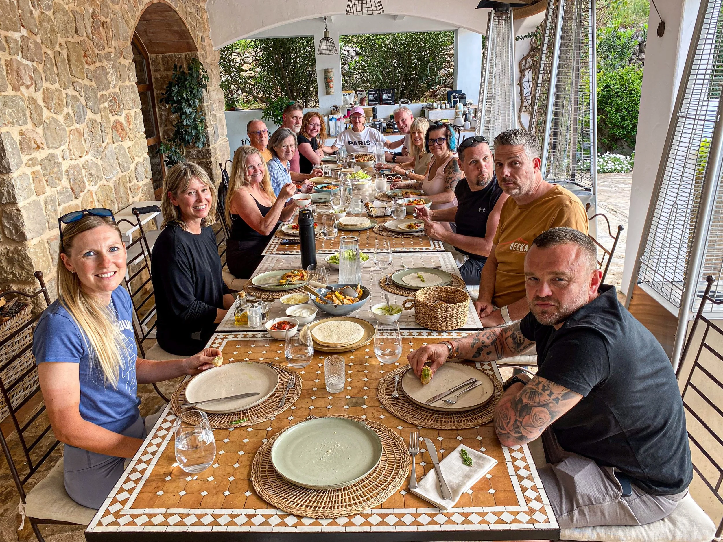 Group of people sitting around a long dining table outdoors, enjoying a meal with plates, bowls, and drinks, in a patio area with stone wall and greenery.
