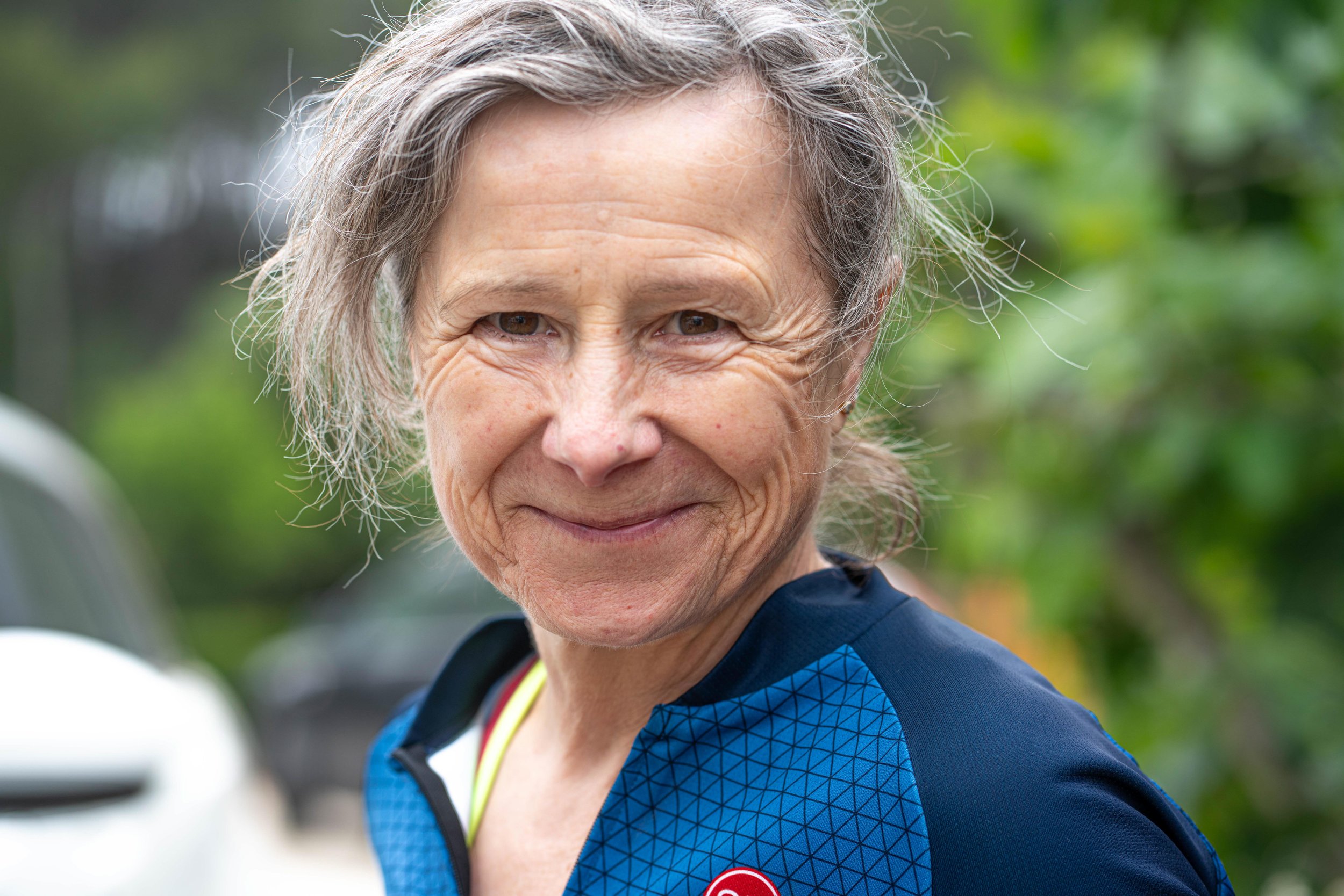 An elderly woman wearing a blue athletic jacket outdoors, smiling at the camera.