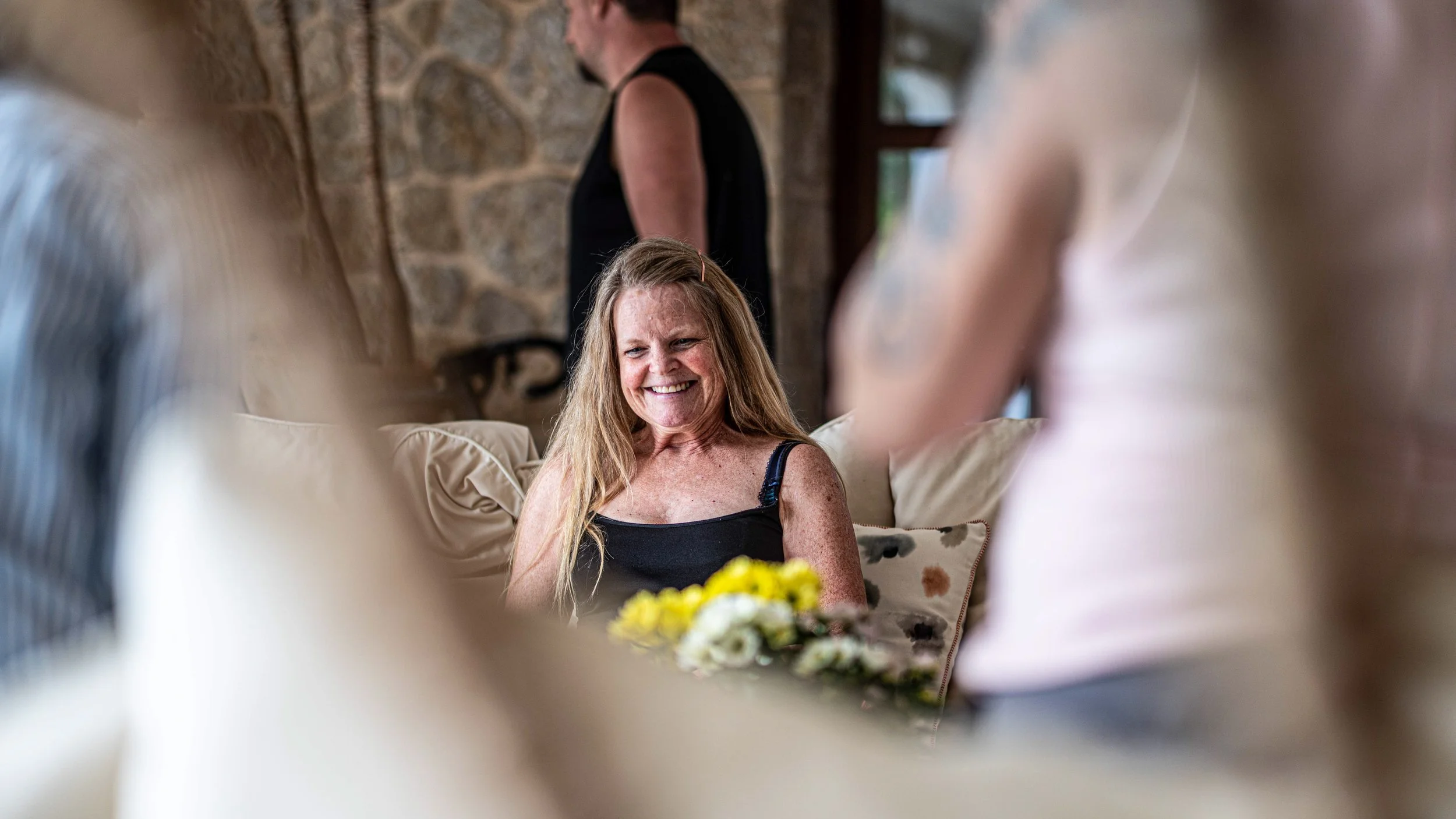 A woman with long red hair, wearing a black tank top, smiling while sitting on a beige sofa surrounded by blurred figures of other women. She is holding a bouquet of yellow and white flowers, and the background features a stone wall.