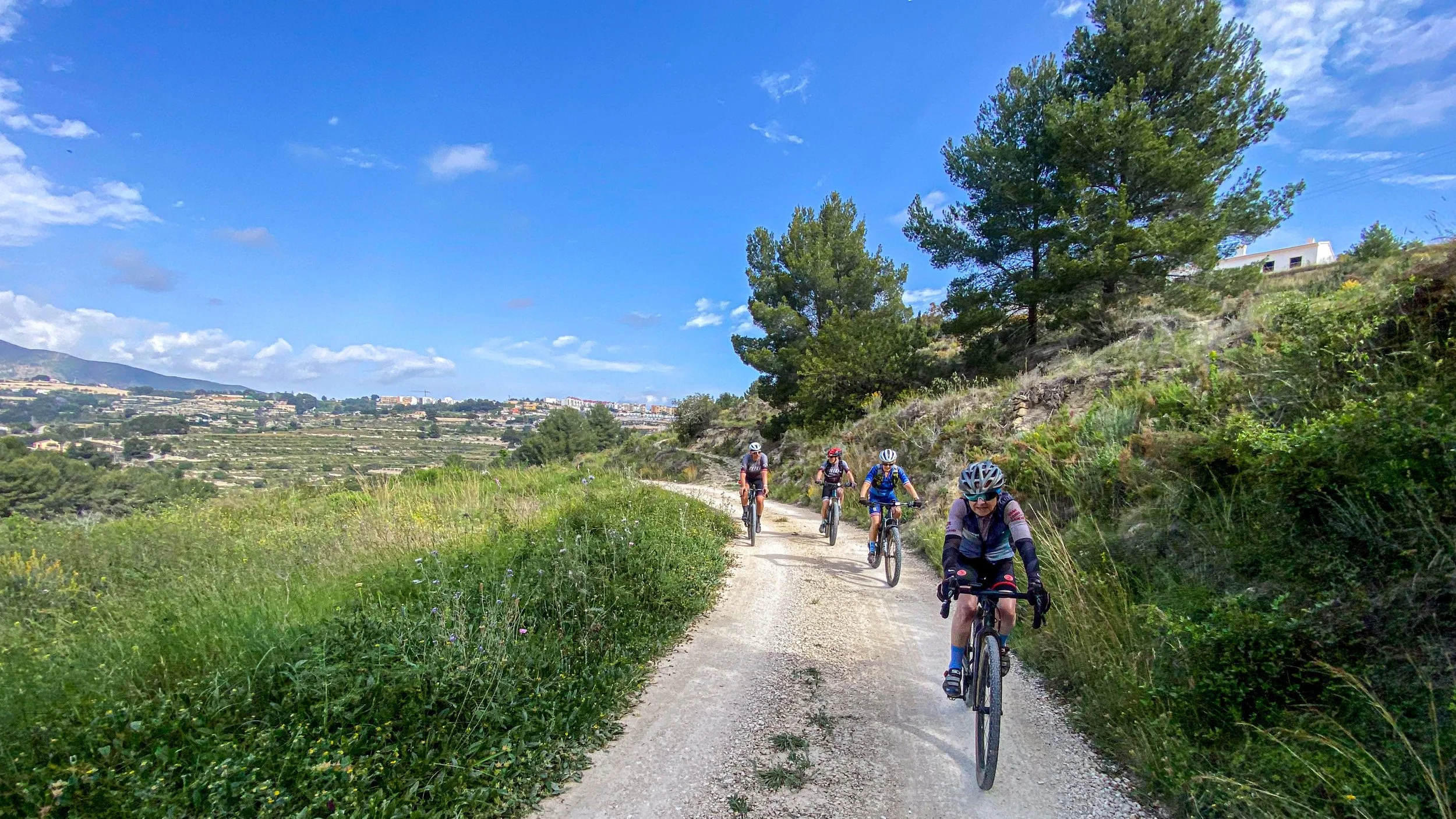 Four cyclists riding on a dirt trail through a green landscape under a blue sky with clouds, surrounded by trees and hills.