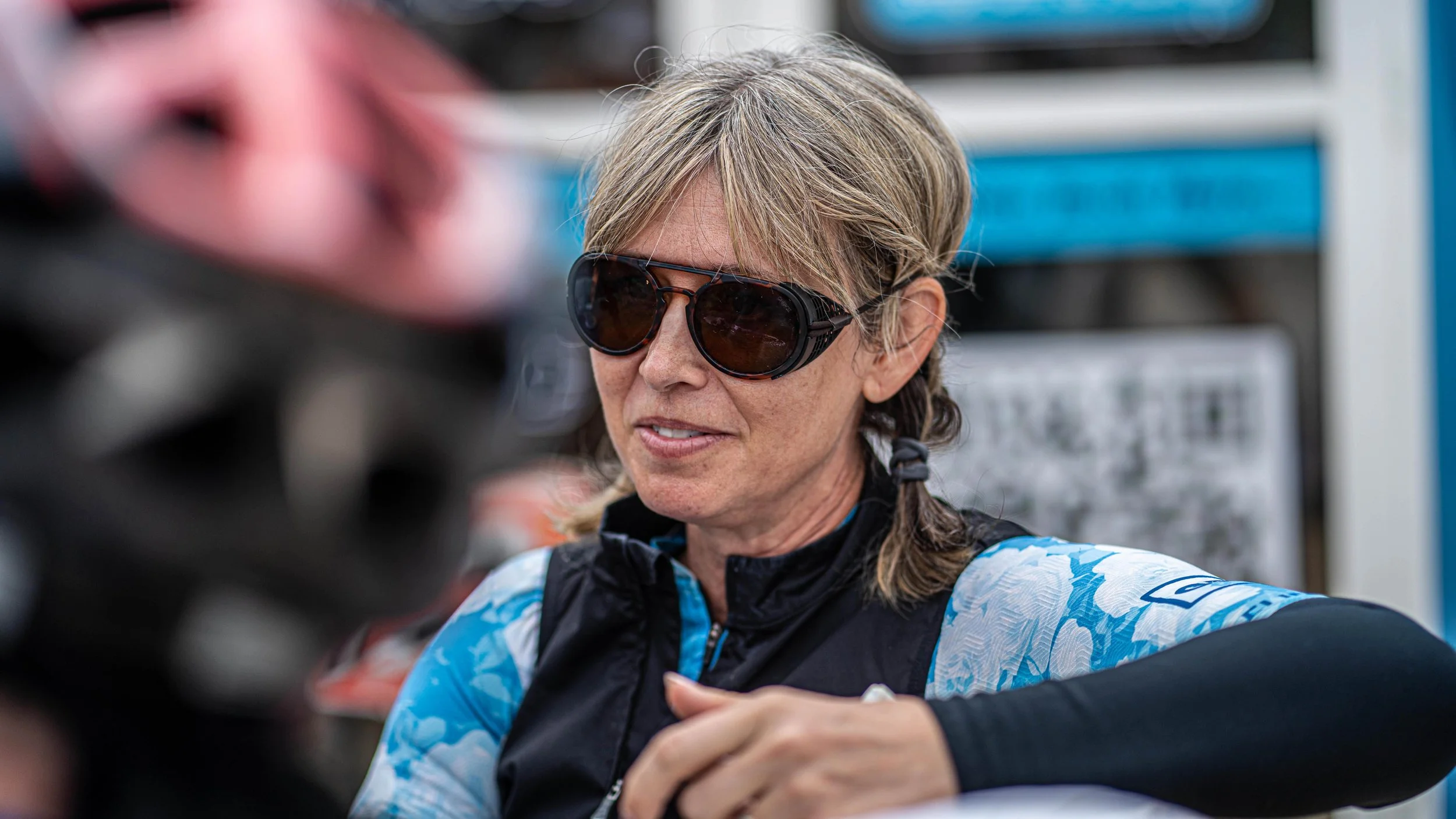 A woman with blonde hair wearing sunglasses and a black and blue athletic jacket, taking part in an outdoor activity.