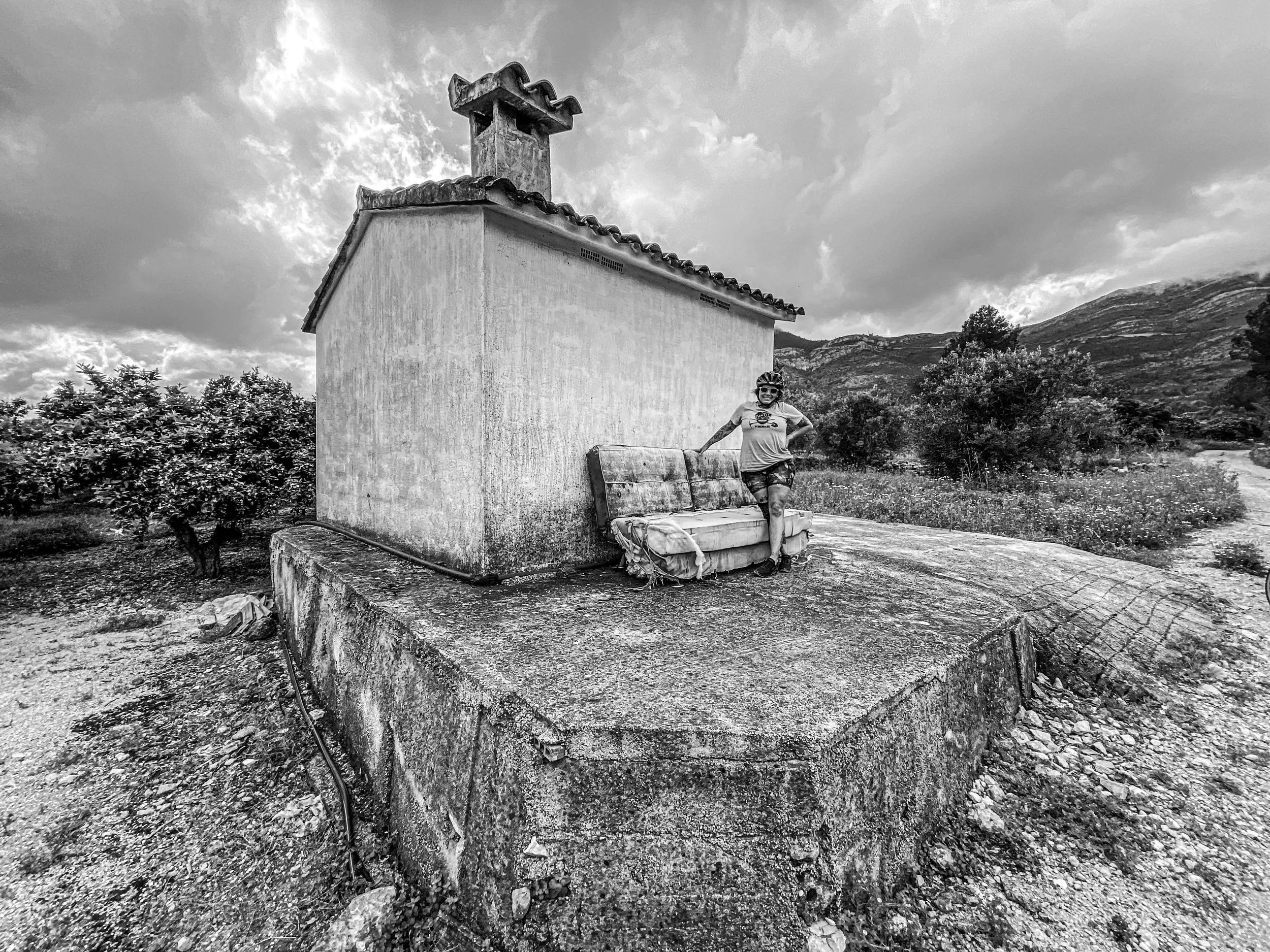 A woman wearing a helmet, t-shirt, and shorts standing next to a worn-out sofa on a concrete platform outside a small building with a chimney, surrounded by trees and mountains, under cloudy sky.