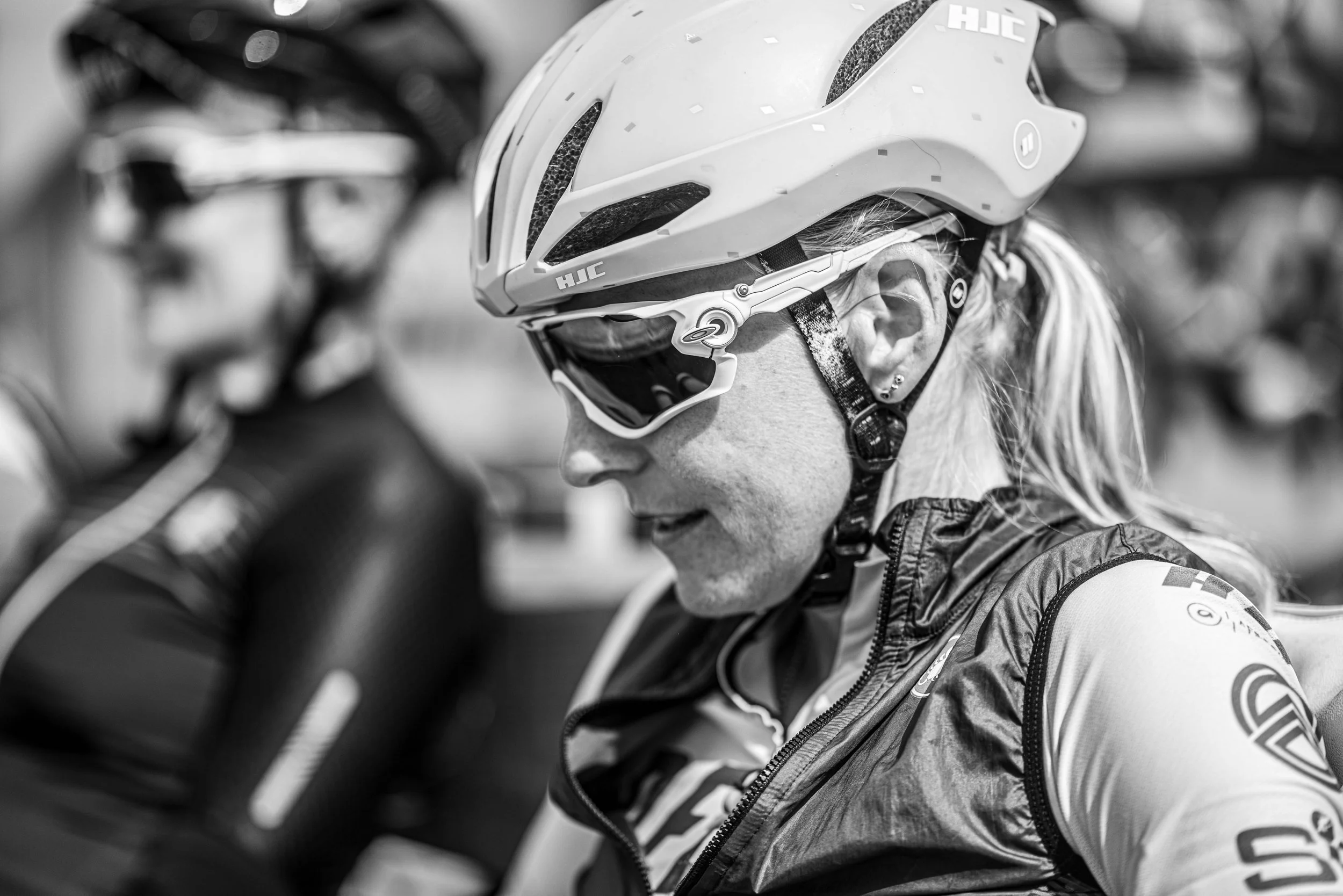 A woman wearing a helmet, sunglasses, and a cycling jersey, preparing for a bike ride, with other cyclists blurred in the background.