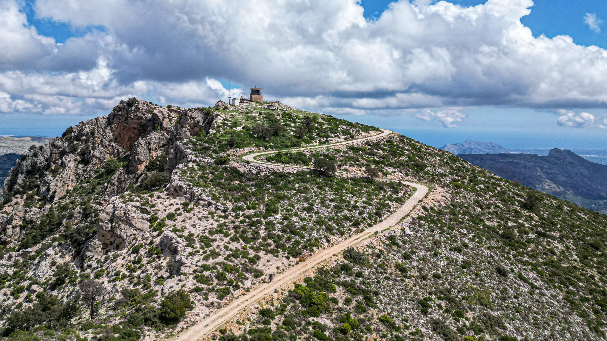 A mountain with a winding dirt road leading to a structure at the top, under a partly cloudy sky.