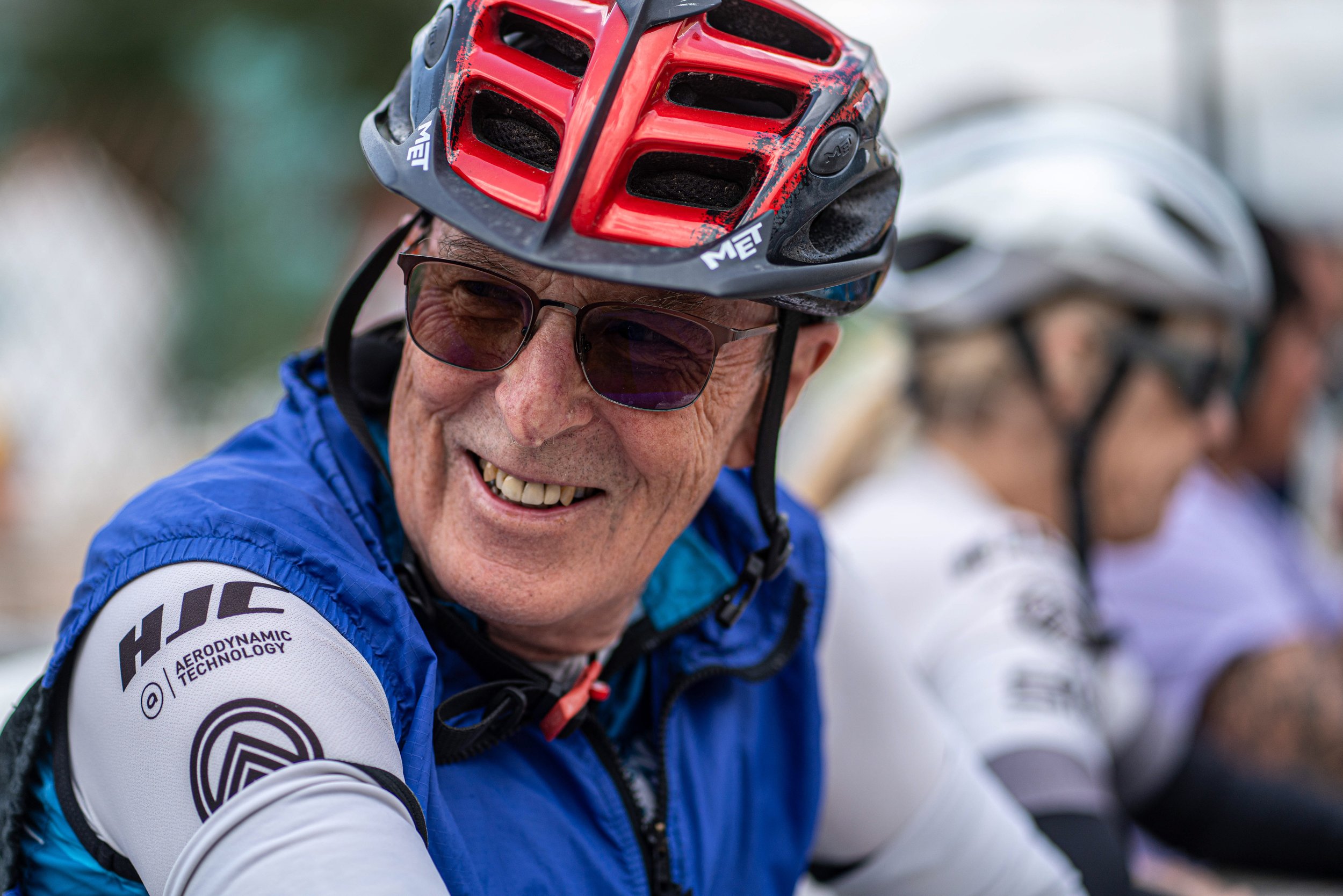 A smiling elderly man wearing a red and black bike helmet, sunglasses, and a blue cycling jersey with white sleeves, participating in a group cycling event with others in the background.