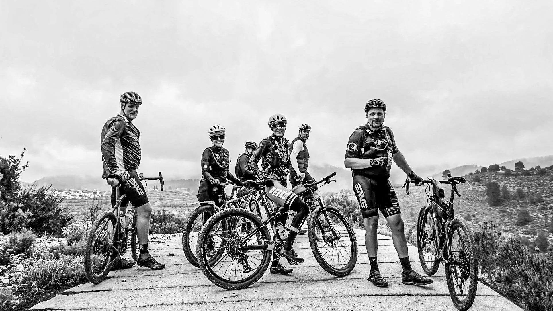 A group of five mountain bikers in cycling gear and helmets, with bicycles, standing on a stone path outdoors with hilly terrain and cloudy sky in the background.