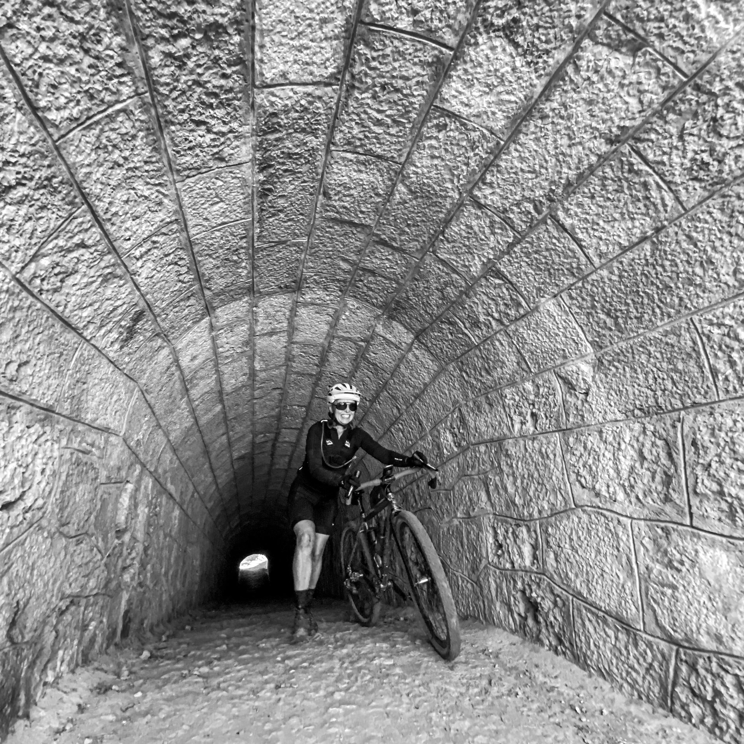 A person with a bicycle standing inside a stone tunnel, smiling and wearing a helmet and sunglasses.