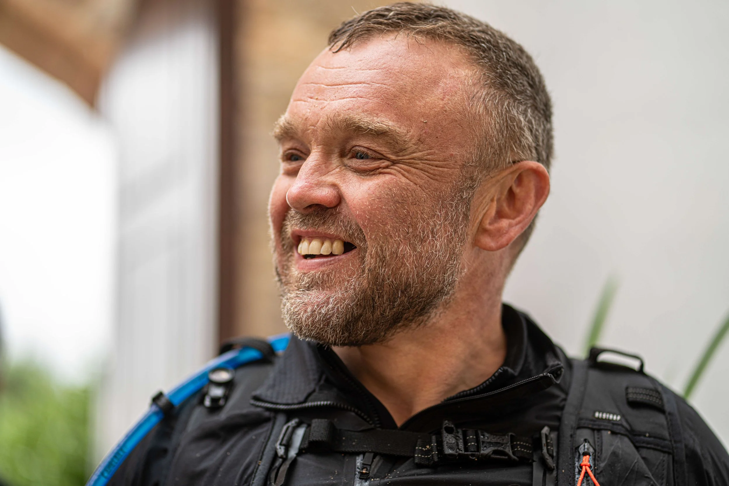 A smiling middle-aged man with a beard, wearing a black jacket with straps, engaging in a conversation outdoors.