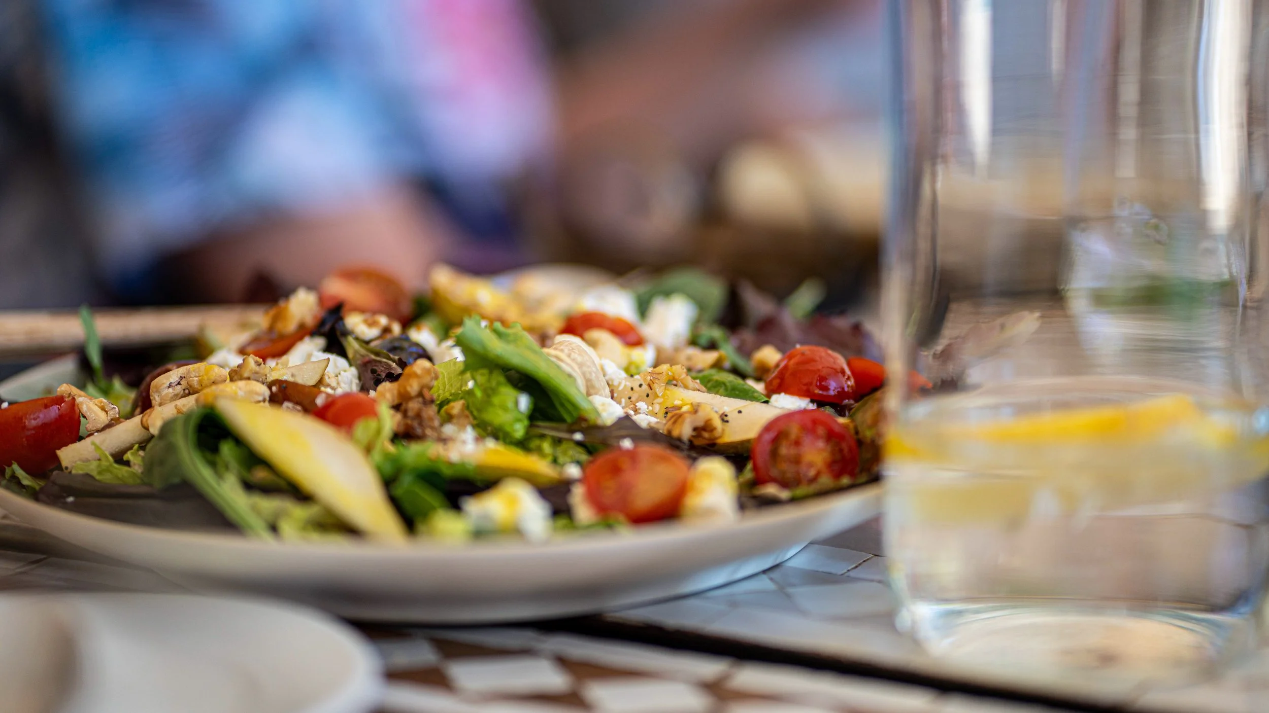 Plate of assorted salad with cherry tomatoes, cheese, greens, and toppings, alongside a glass of water with lemon slices.