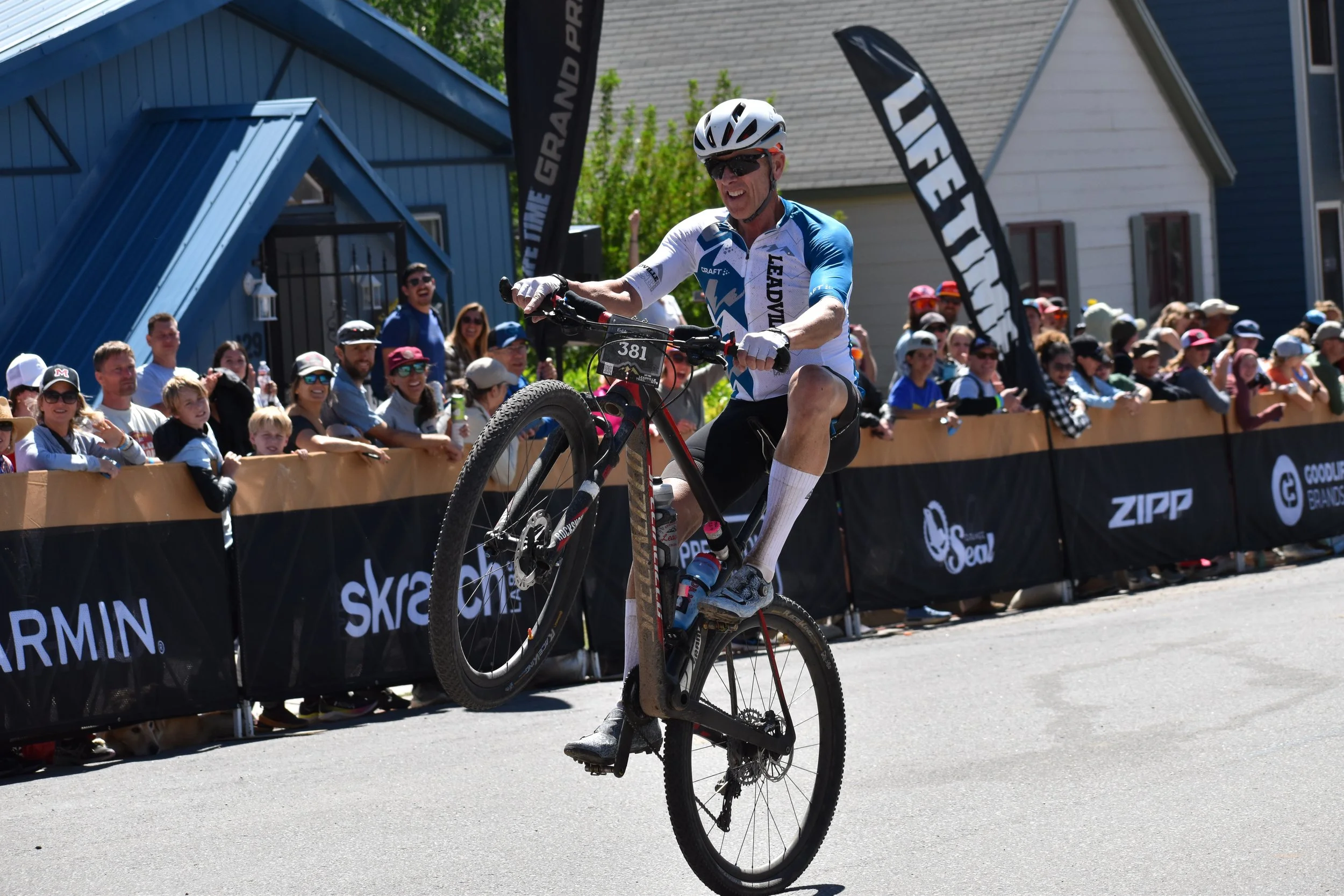 A cyclist performing a wheelie on a mountain bike during a race, with cheering spectators behind the barriers on a sunny day.