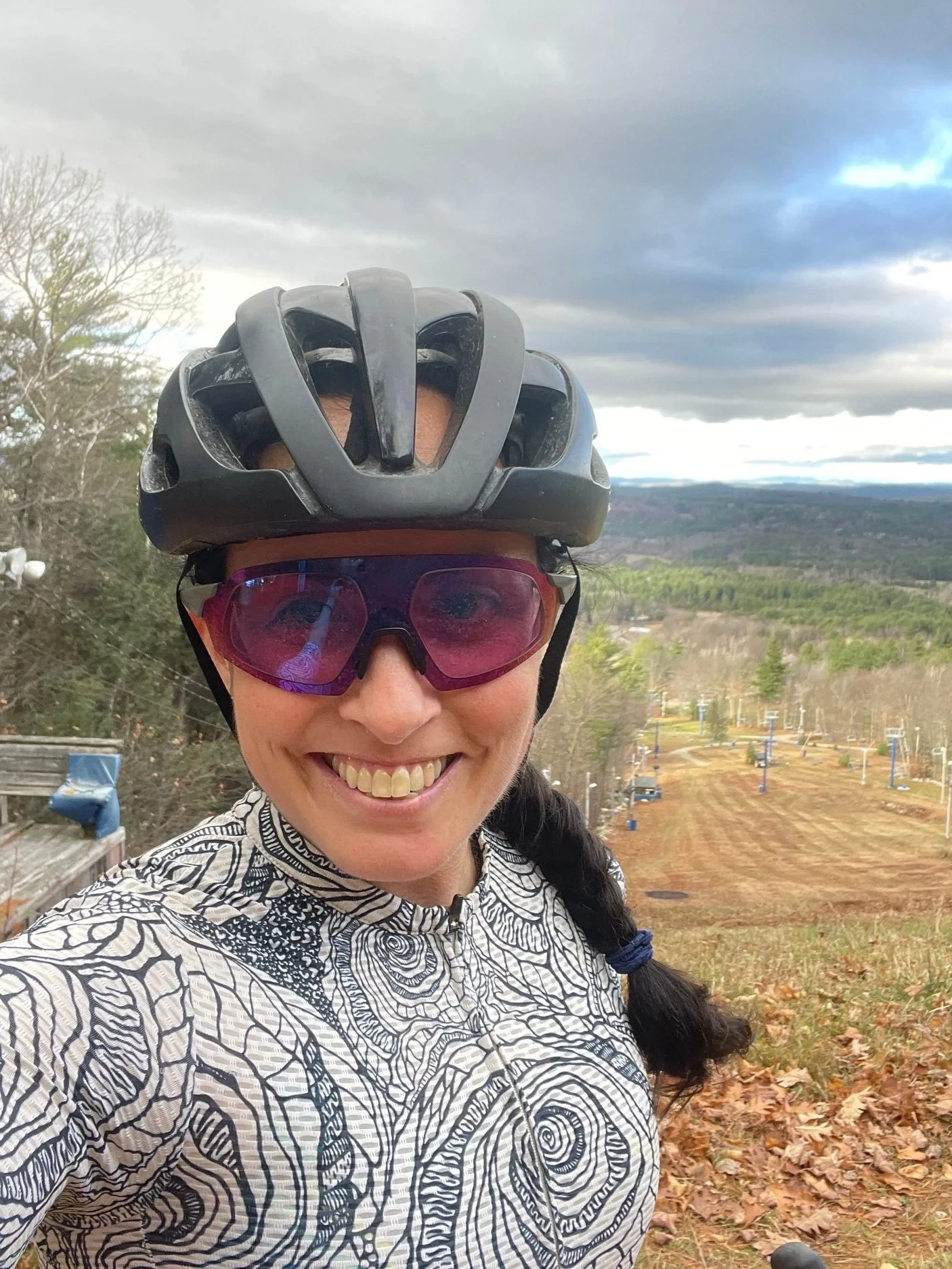 Smiling woman wearing a bike helmet and sunglasses, taking a selfie outdoors with a background of ski lift chairs and a mountain landscape.