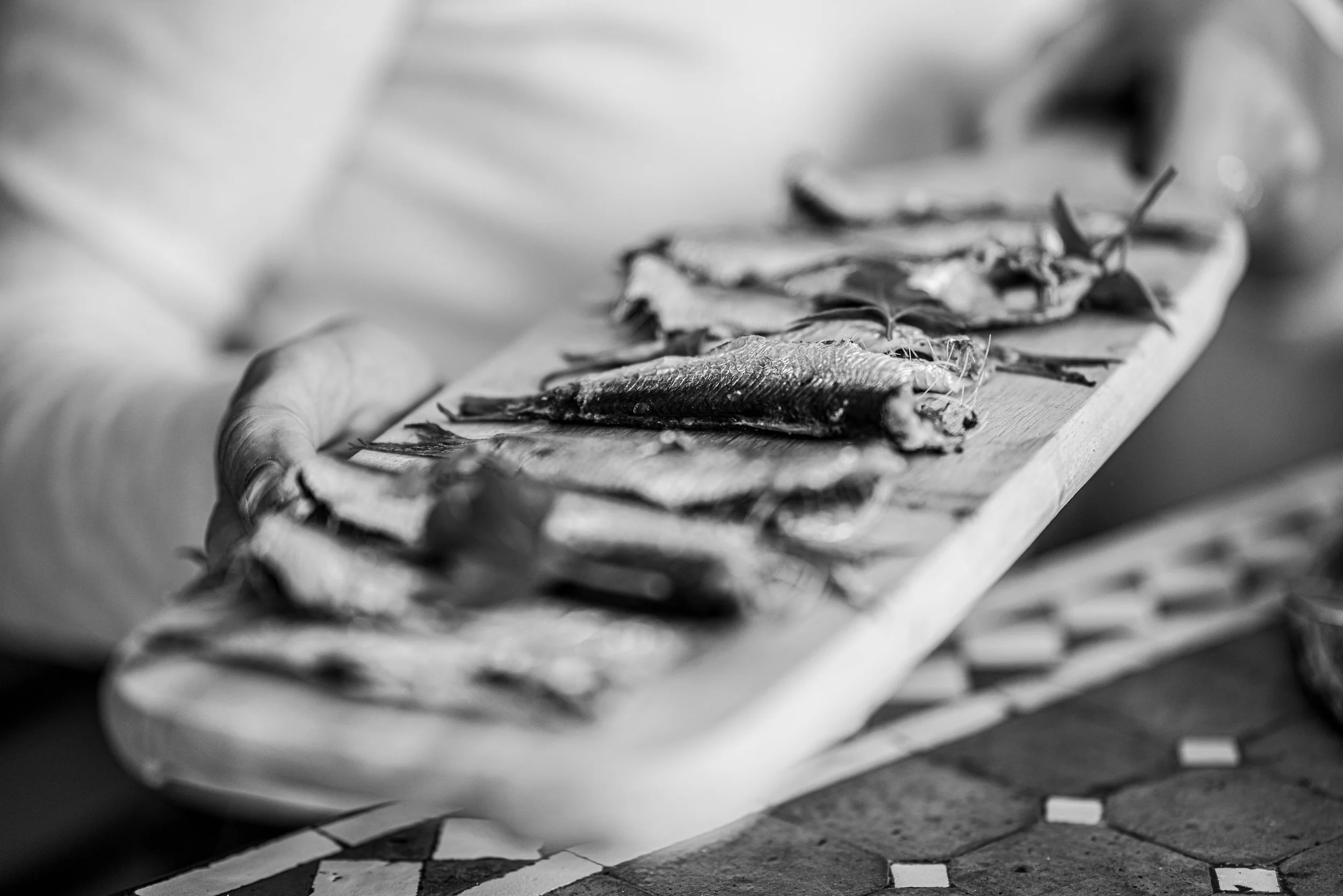 Black and white photo of a person holding a wooden board with dried fish and herbs on top.
