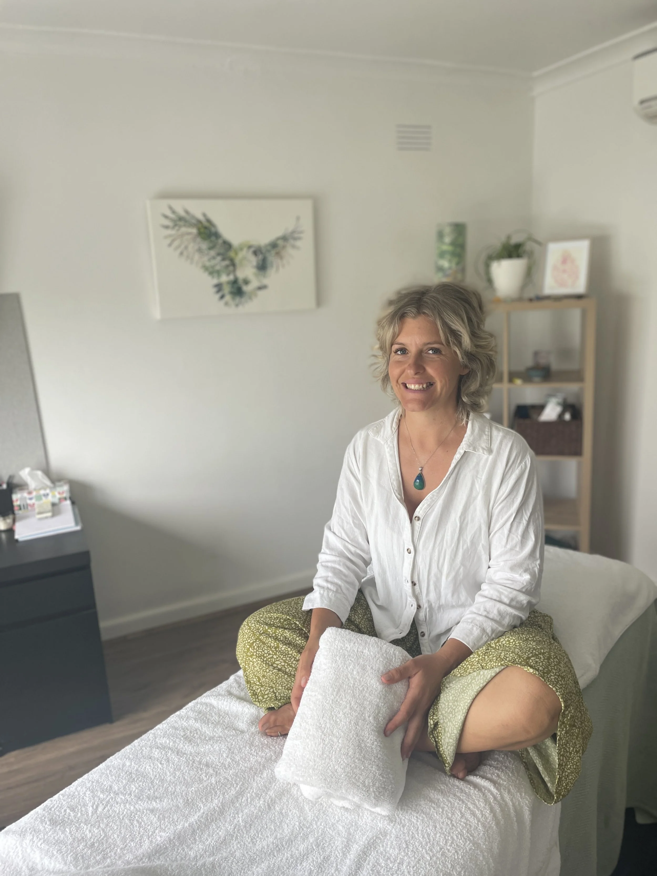 A woman sitting cross-legged on a bed with a white pillow, smiling at the camera, in a room with minimal decor and artwork on the wall.