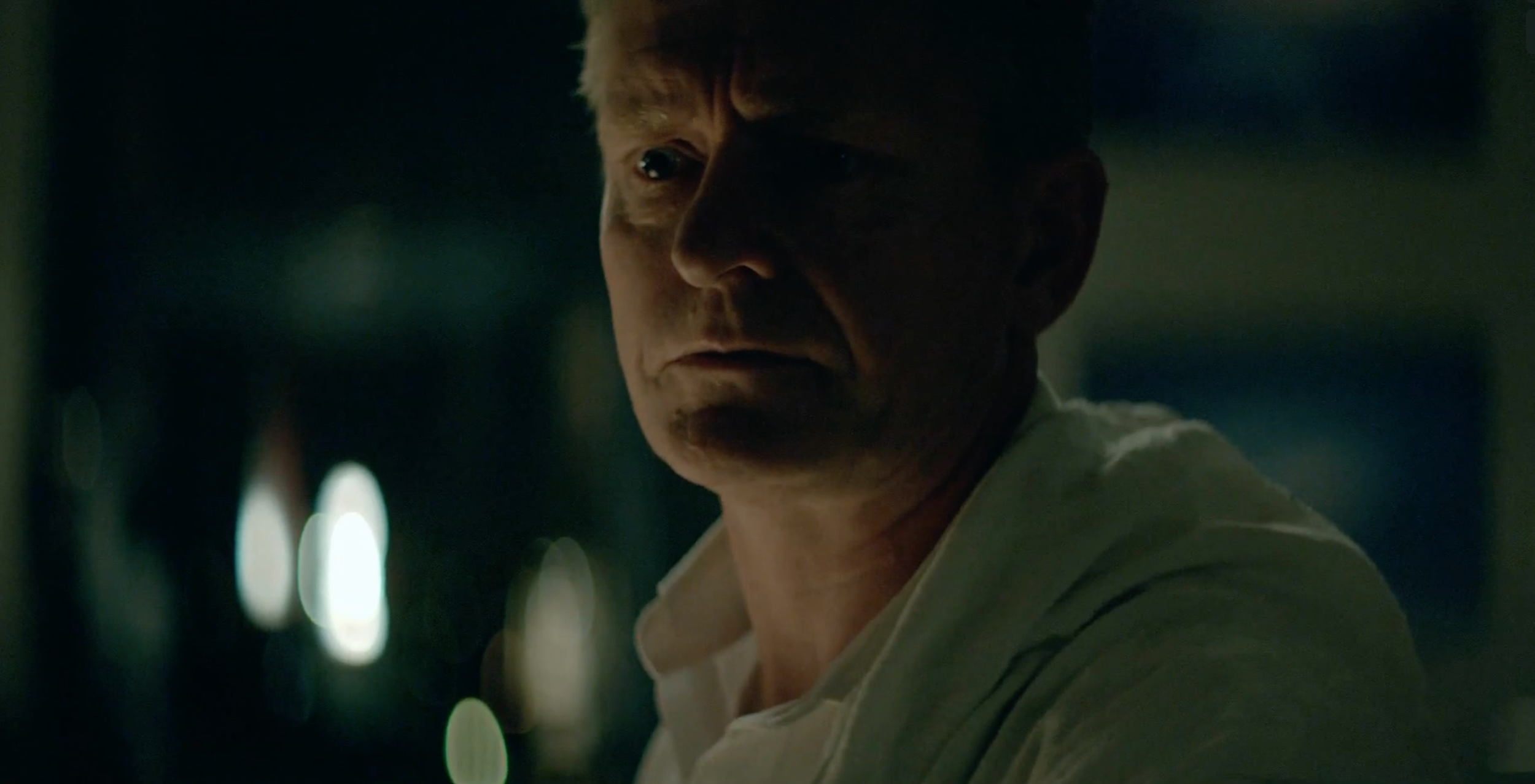 A close-up of a man with short hair in a dimly lit room, with a serious expression, facing slightly to the right. occupied tv series.
