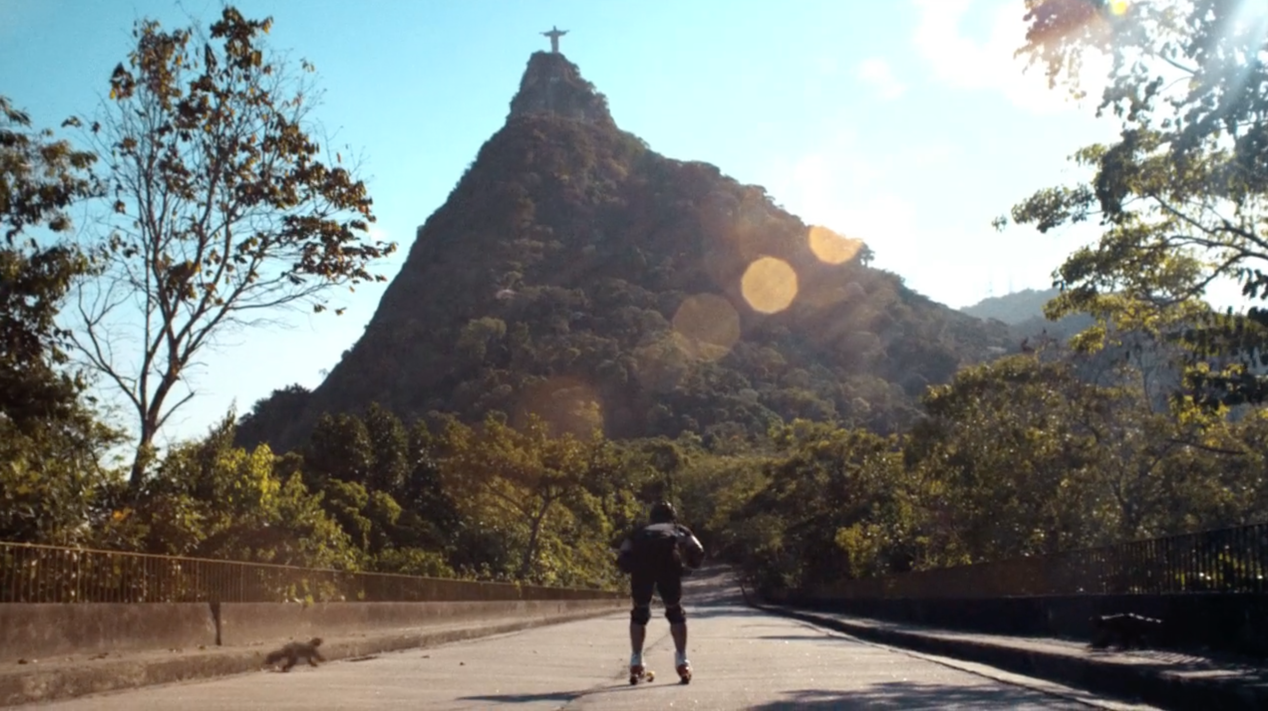 A person rollerblading on a path in a park with a view of the Christ the Redeemer statue on the top of a mountain in the background, surrounded by trees and sunlight. lillyhammer tv series - netflix
