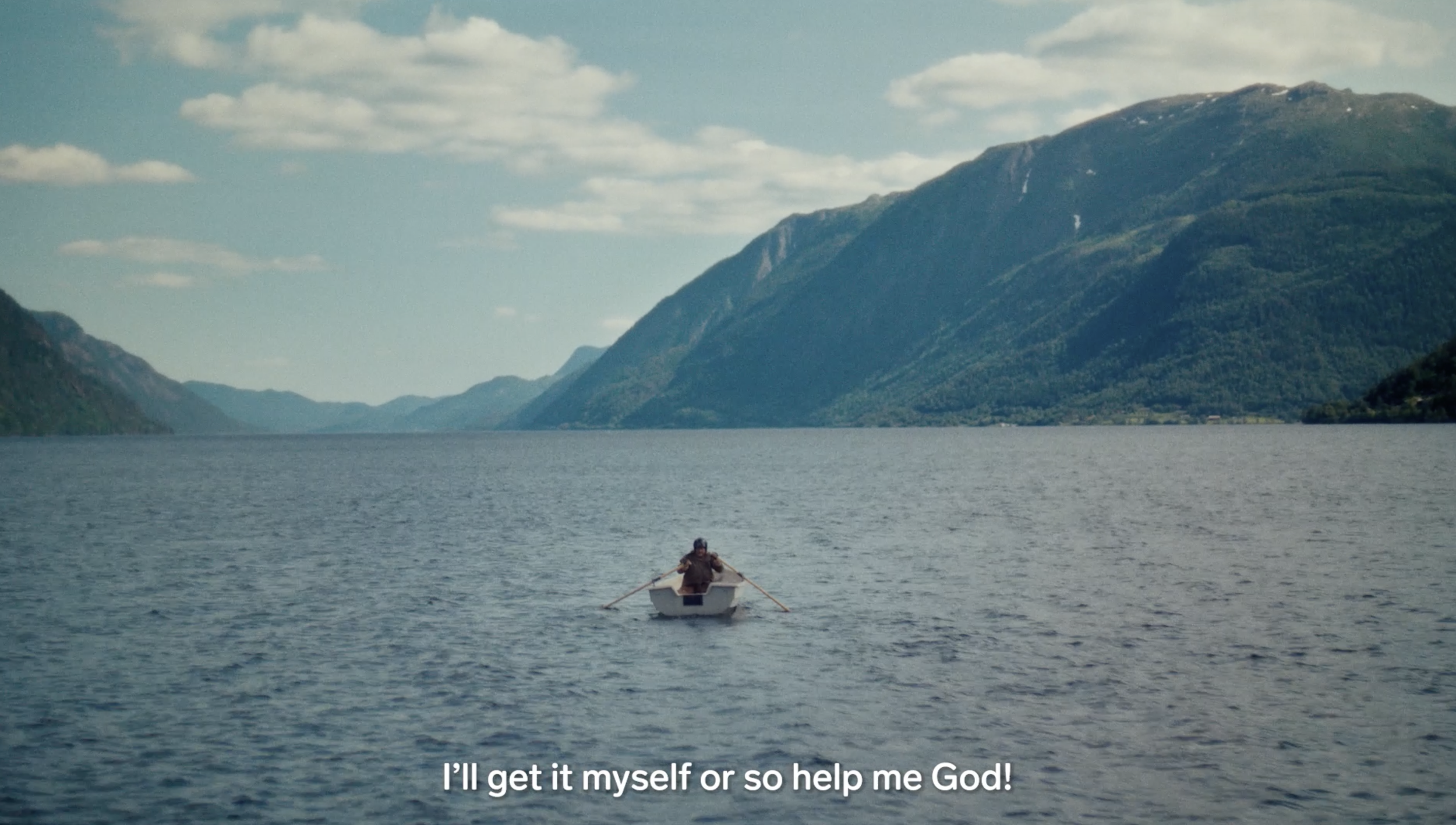 A person rowing a small boat on a large body of water, surrounded by mountains with a partly cloudy sky overhead. finn commercial