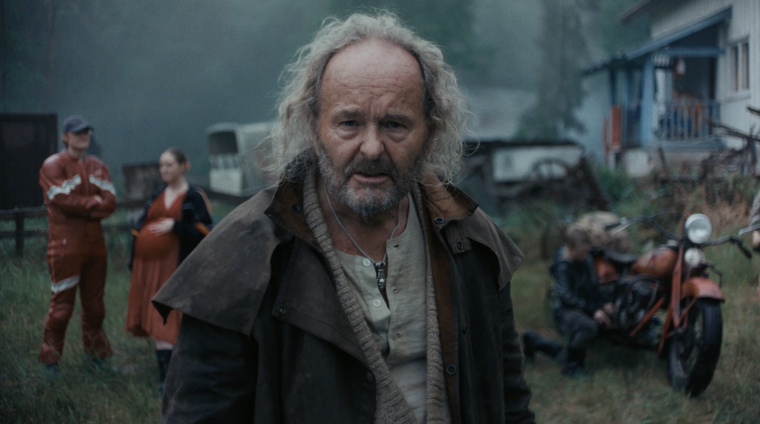 Finn commercial. A bearded elderly man with curly hair and a serious expression, wearing a brown jacket, stands outdoors with a group of blurred people and motorcycles in the background.