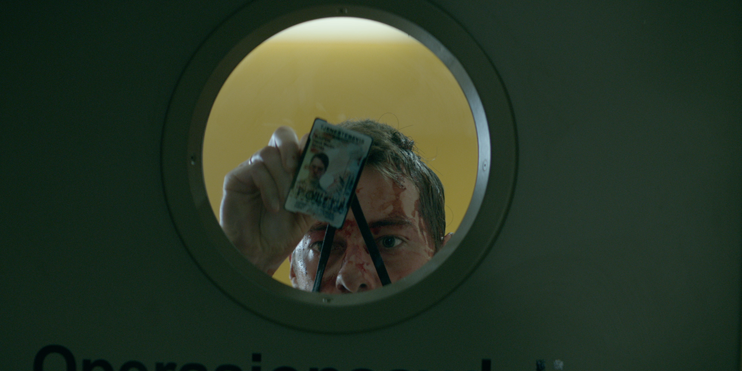 A person with blood on their face looking into a circular opening, holding an ID card toward the camera. occupied tv series.