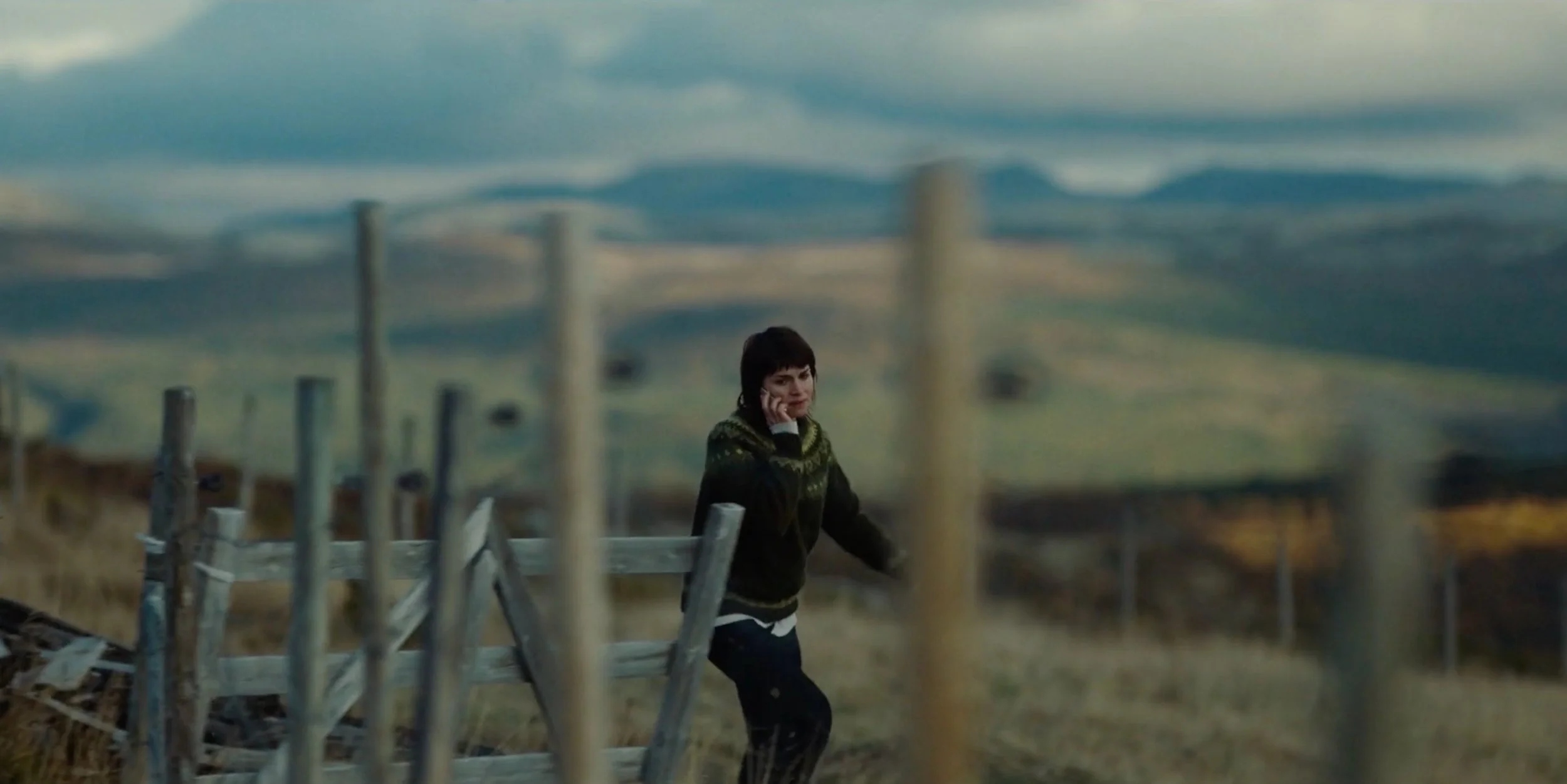 A woman talking on a cellphone outdoors near a fence, with rolling hills and a cloudy sky in the background. fenris tv series