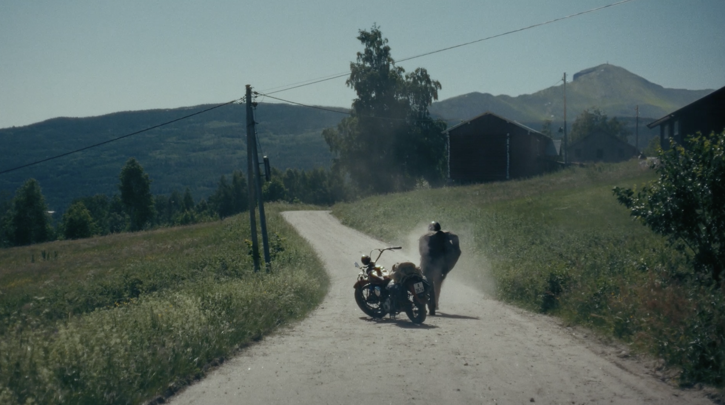 A motorcycle on a dirt road with a rider walking nearby, set in a rural landscape with grass, trees, houses, and mountains in the background. Finn commercial.