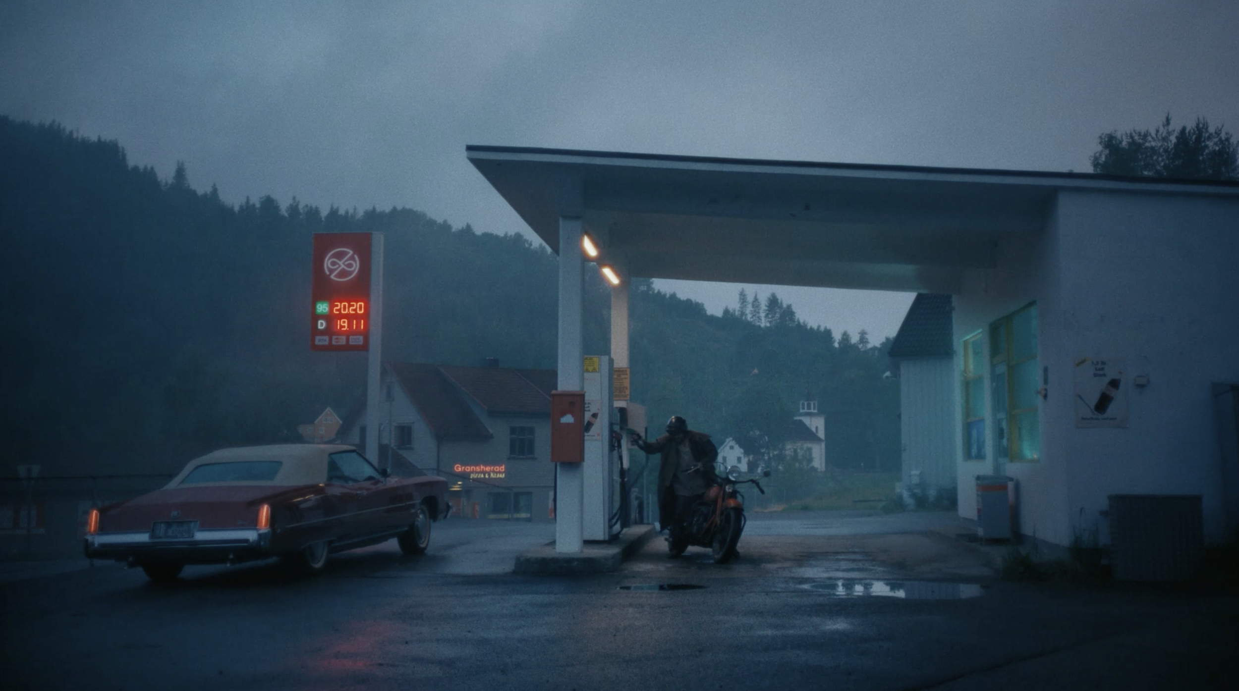A gas station with a person refueling a motorcycle during dusk, with a car parked nearby and a sign displaying fuel prices and type. finn commercial