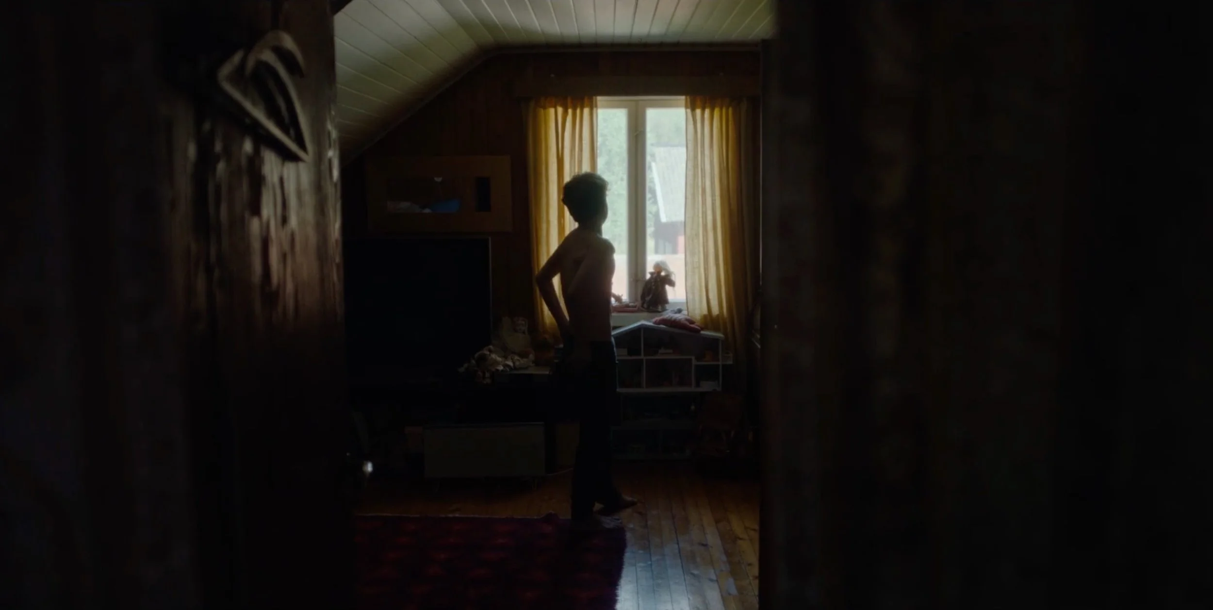 A person standing in a dimly lit room by a window with yellow curtains, with sunlight coming in, wooden floor, and furniture. fenris tv series