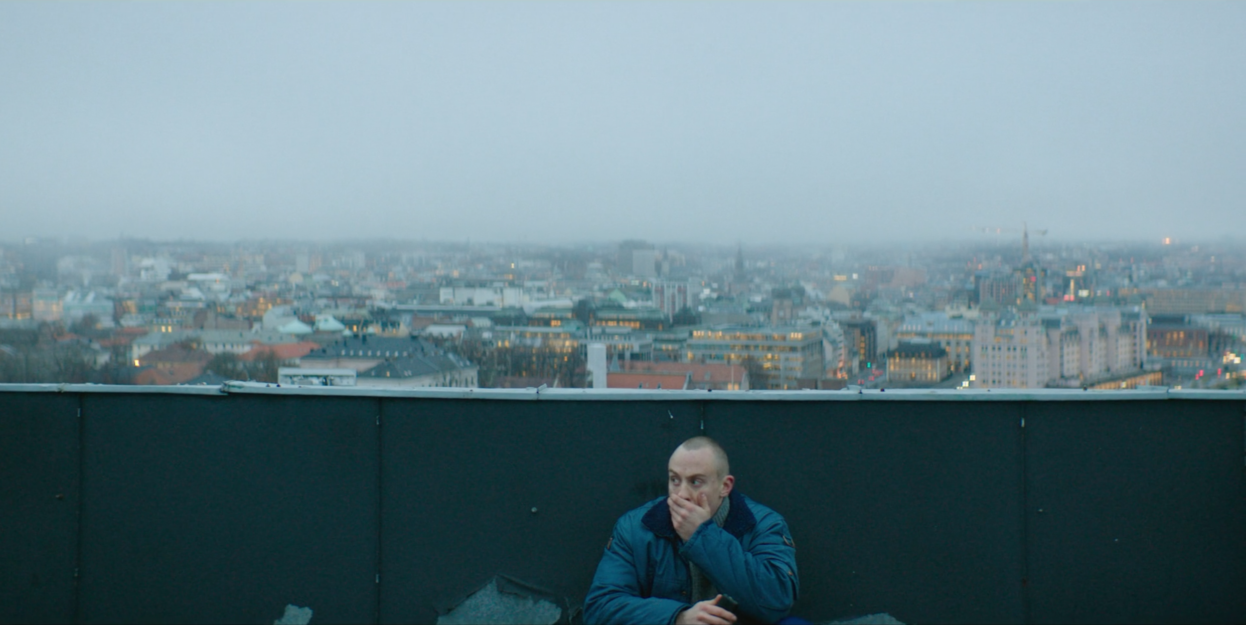 A man in a blue jacket sitting on a rooftop ledge with a city skyline in the background, covering his mouth with one hand. valkyrien tv sereis