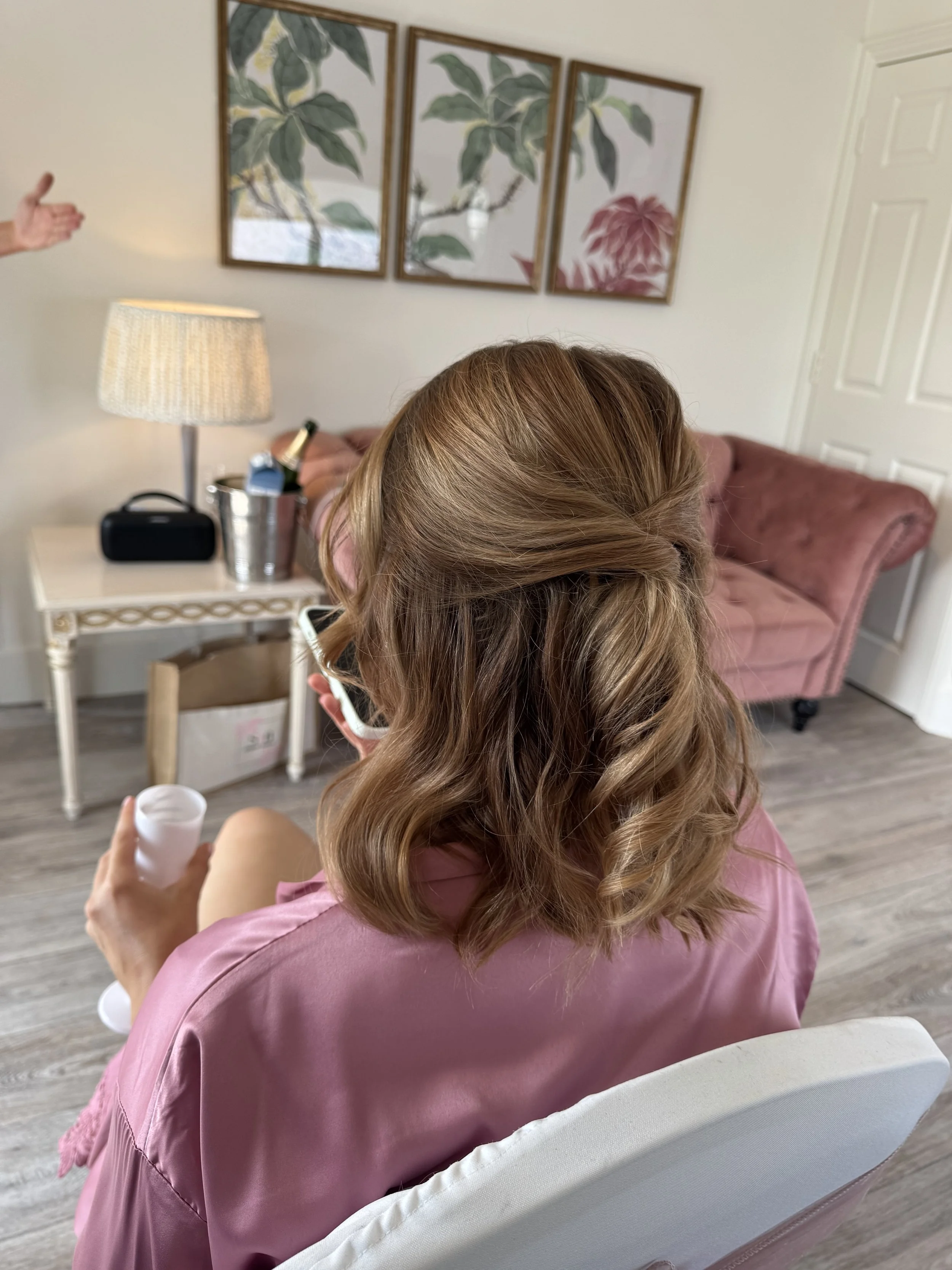 A woman with long, curly, light brown hair is sitting on a white chair, wearing a pink satin robe. She is holding a small cup in her left hand while looking at her phone. Behind her, there is a white desk with a lamp, a small black speaker, a silver ice bucket with a bottle, and some other items. On the wall, there are three framed botanical prints. There is also a pink velvet armchair in the background.