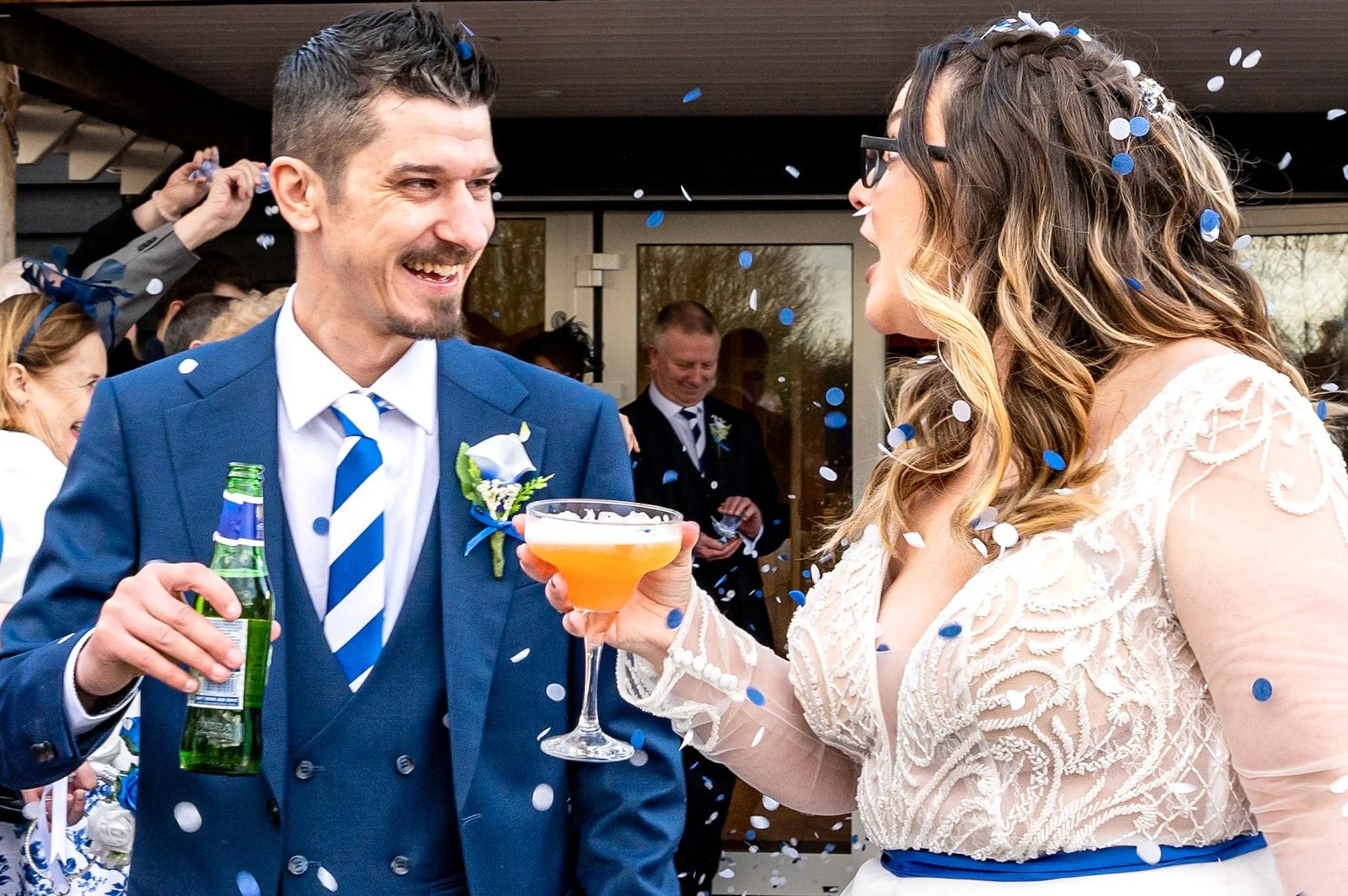 A bride and groom celebrating with confetti at their wedding. The bride is holding a cocktail glass, and the groom is holding a beer bottle. They are smiling and looking at each other.