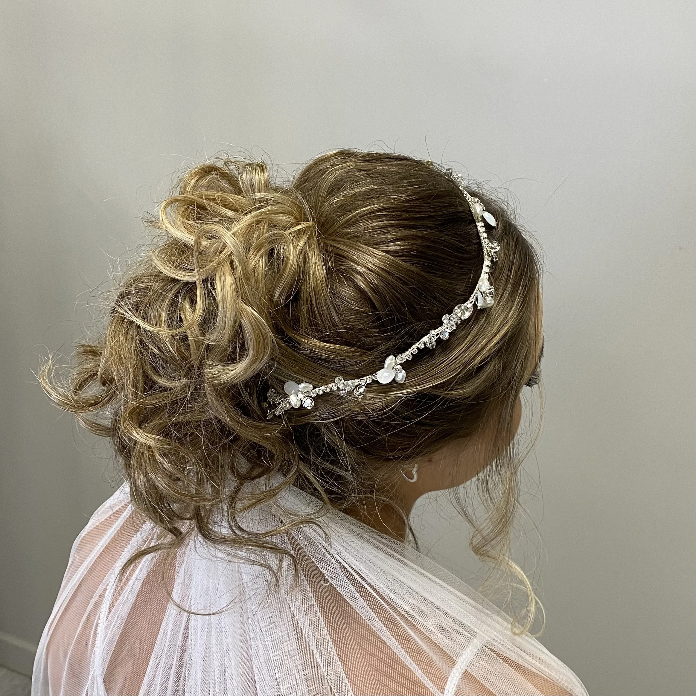 Close-up of a woman with curly blonde hair styled in an updo, adorned with a pearl and rhinestone hair accessory, wearing a light-colored tulle dress.