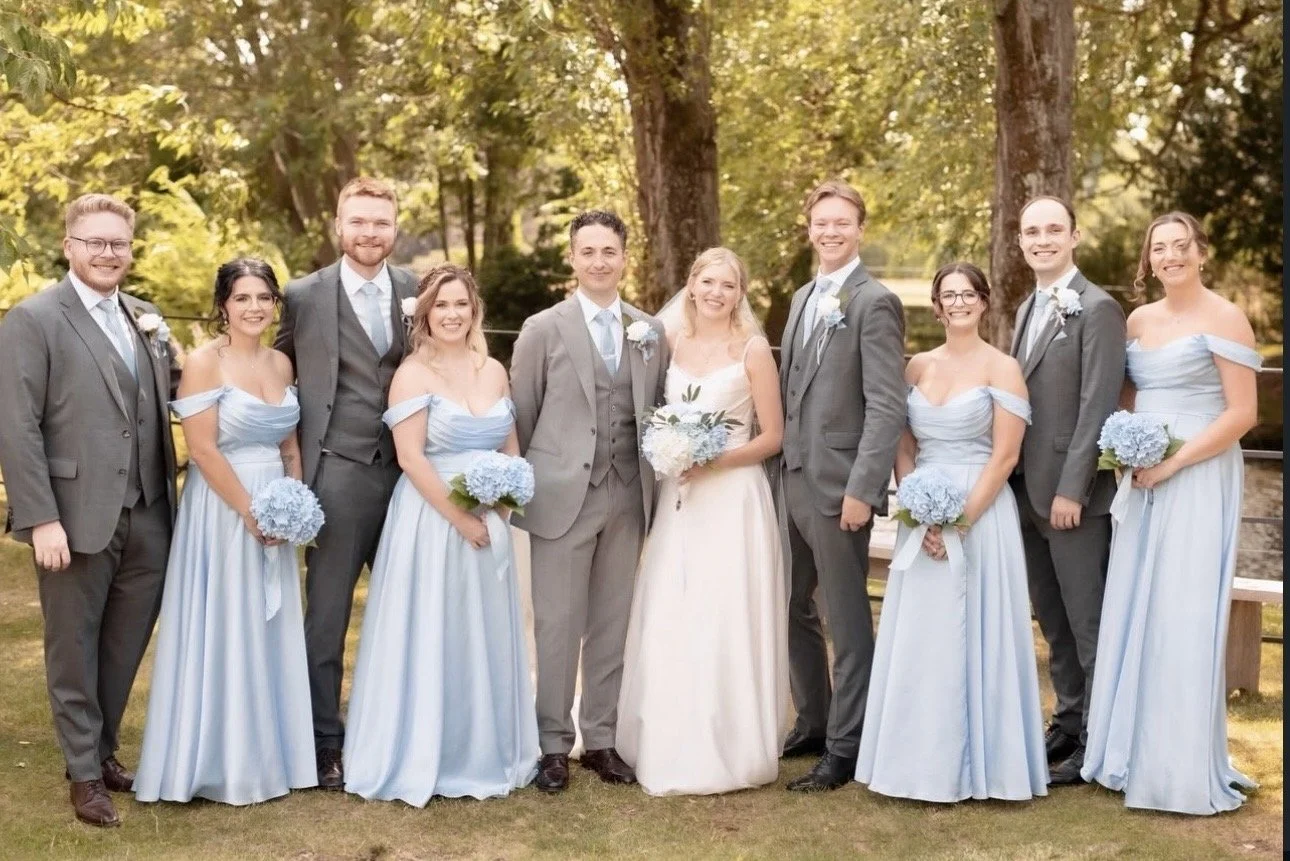 Group of people in wedding attire standing outdoors in a park with trees. The bride is in the center in a white wedding dress holding a bouquet, surrounded by bridesmaids in light blue dresses and groomsmen in gray suits with light blue ties, all smiling.