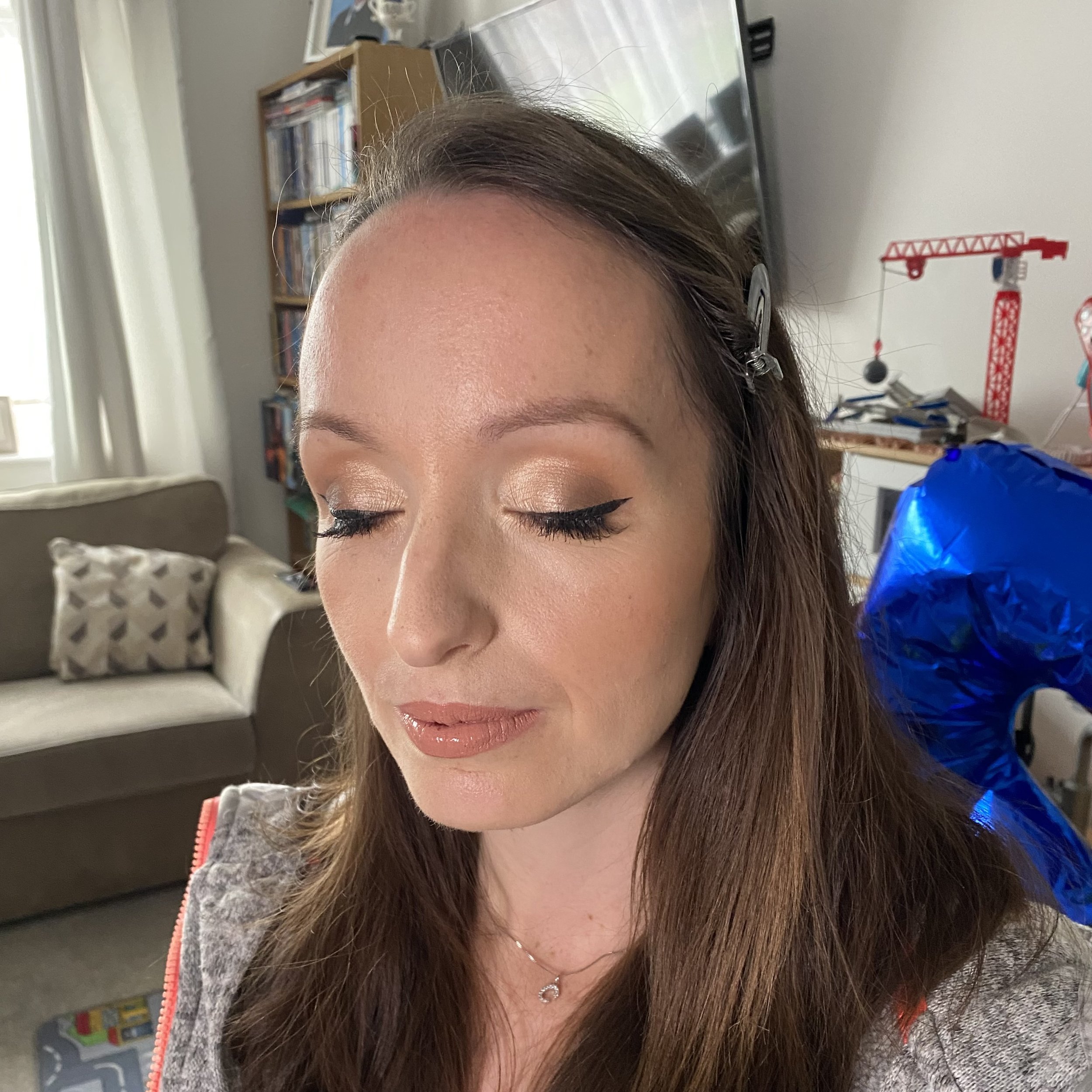 A close-up of a woman's face with closed eyes, brown hair, wearing makeup, with background elements including a bookshelf, a red crane toy, and a window with curtains.