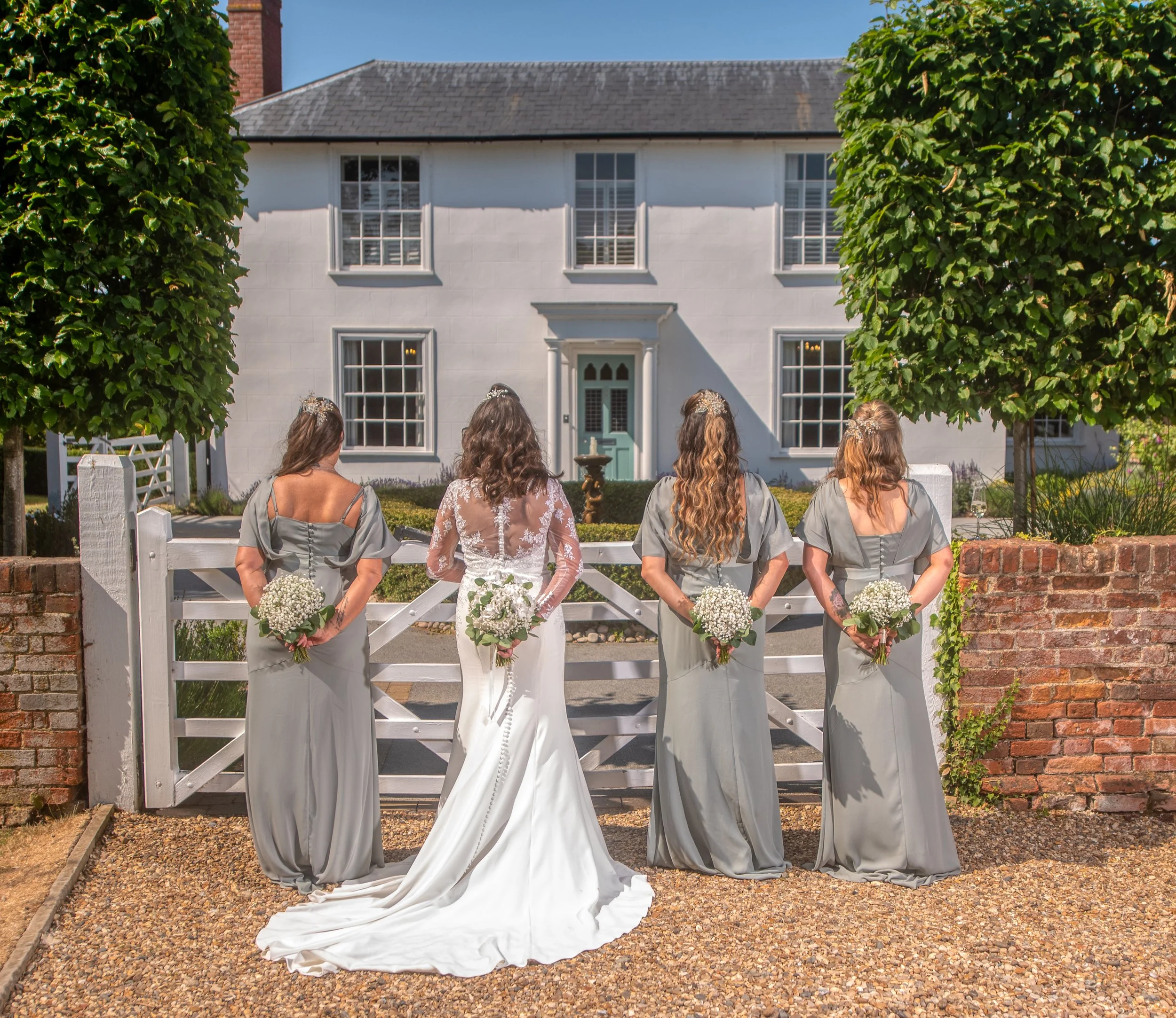 Four women in lavender and gray dresses standing in front of a white house with a fountain, holding bouquets of white flowers, during a sunny day.