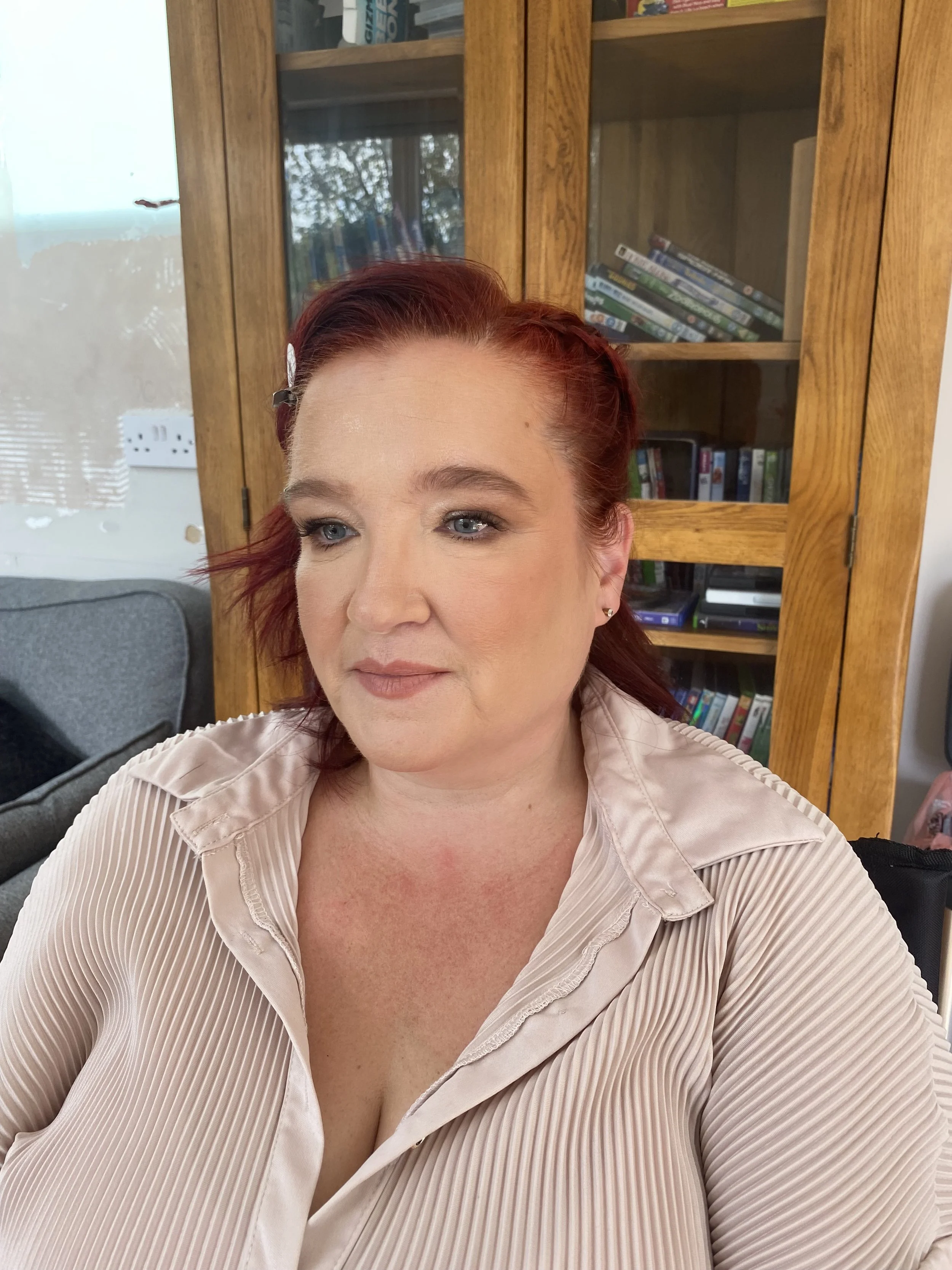 A woman with red hair and blue eyes sitting indoors in front of a wooden bookshelf filled with DVDs and books, wearing a beige pleated blouse.