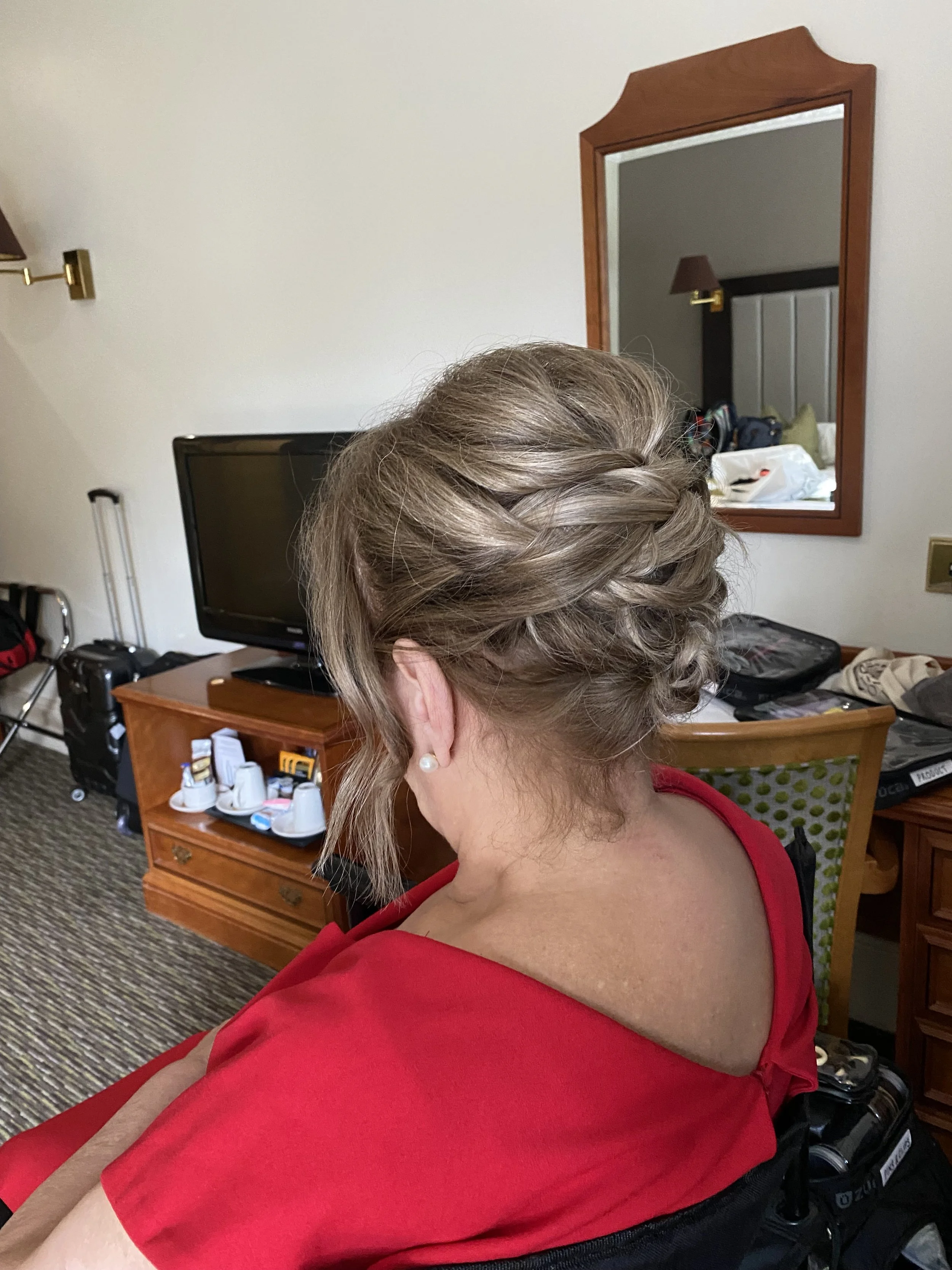 A woman with short, styled hair sitting in a hotel room. She is wearing a red sleeveless top and pearl earrings. In the background, there is a wooden desk with a TV, a small shelf with mugs, a luggage area with suitcases, and a large mirror reflecting a bed, bedside table, and a wall-mounted lamp.