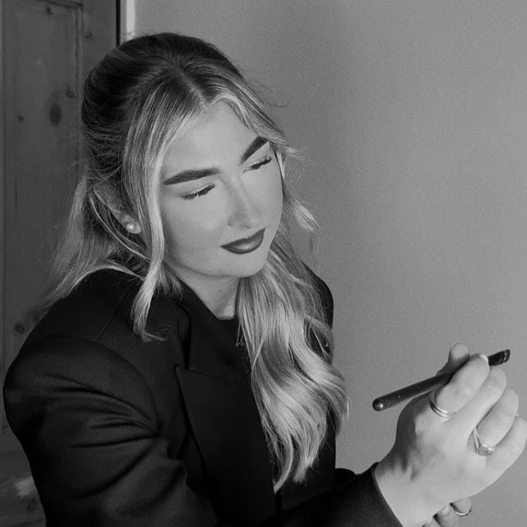 A young woman with long wavy blonde hair, stylish makeup, and earrings, holding a pen or pencil, looking down with a slight smile.