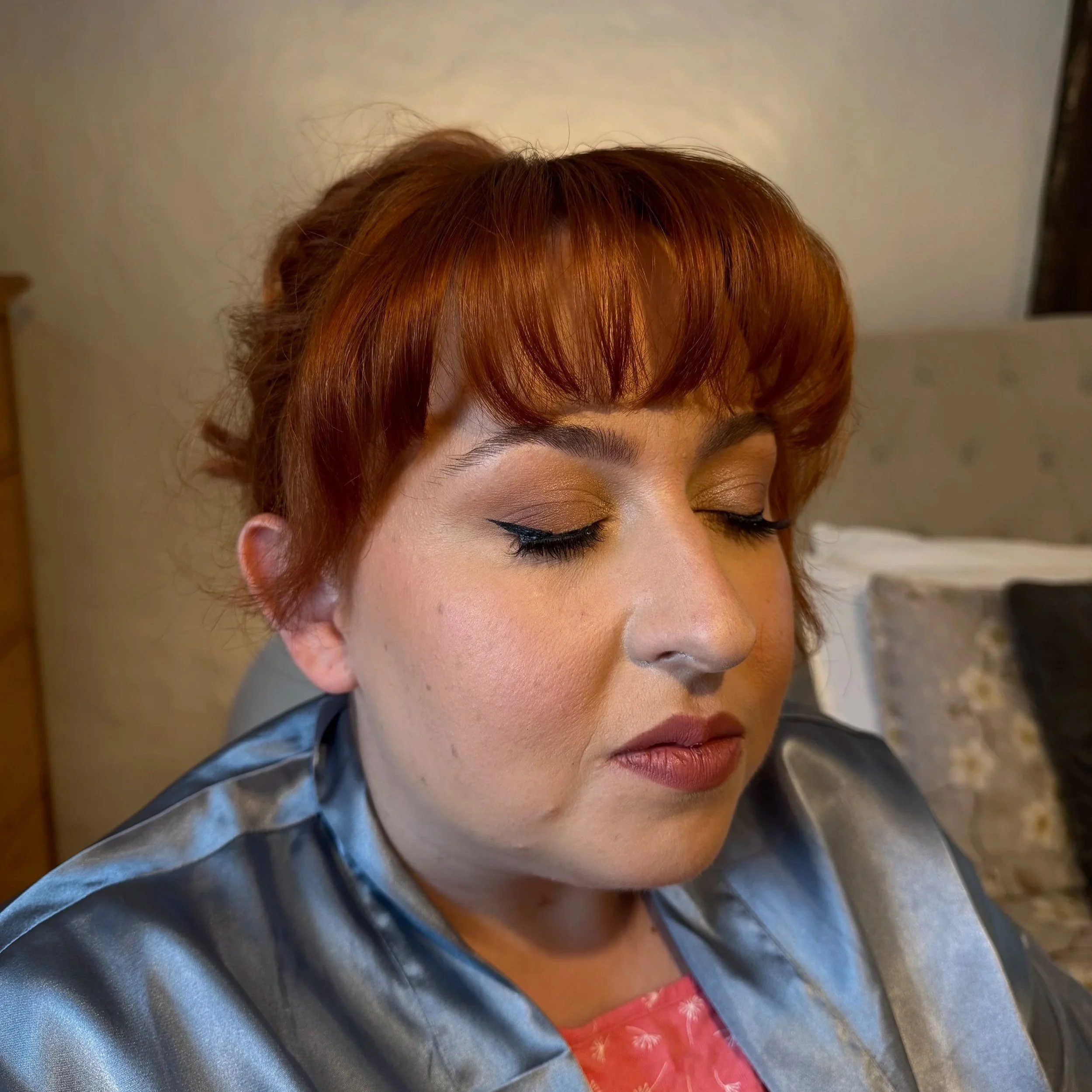 Close-up of a person with short, reddish-brown hair and closed eyes, wearing makeup, a gray jacket, and a red garment with patterns, against a neutral background.