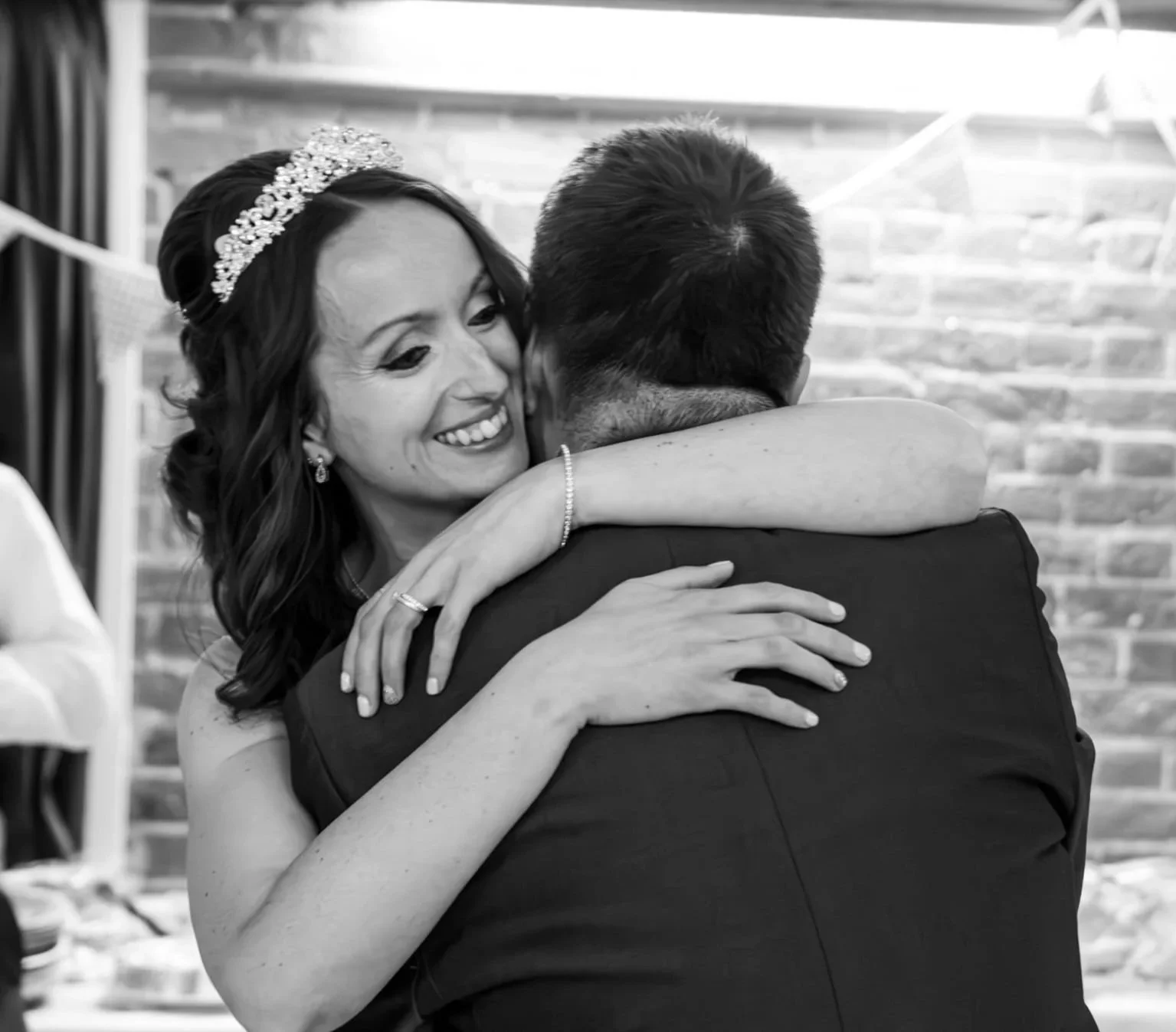 A woman and a man are embracing at a wedding reception. The woman is smiling and wearing a tiara, earrings, and a bracelet. The man is dressed in a suit and facing away from the camera. They are hugging tightly with a brick wall in the background.