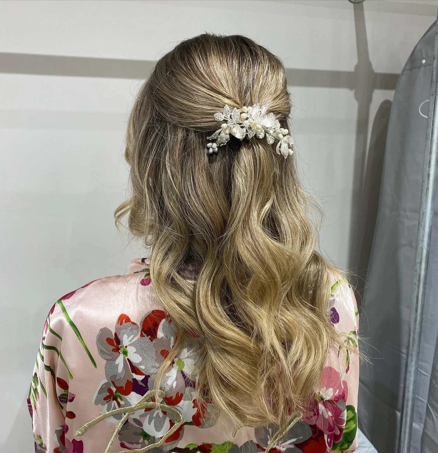 Back view of a woman with long, wavy blonde hair styled with a decorative hairpiece adorned with pearls and crystals, wearing a pink floral kimono-style garment.