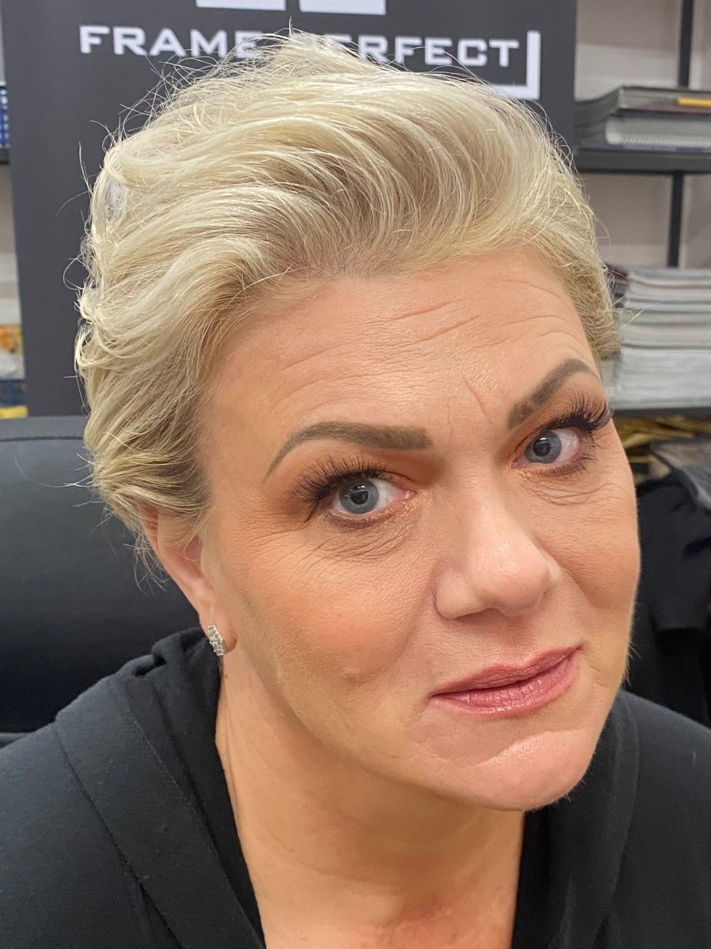 Close-up of a woman with short, blonde hair styled upwards and slightly to the side, with makeup including dark eyeliner, mascara, and lipstick, sitting in an indoor setting with shelves and signage in the background.