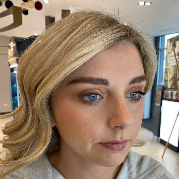 Close-up of a blonde woman with blue eyes and makeup, looking slightly downward, in an indoor setting.