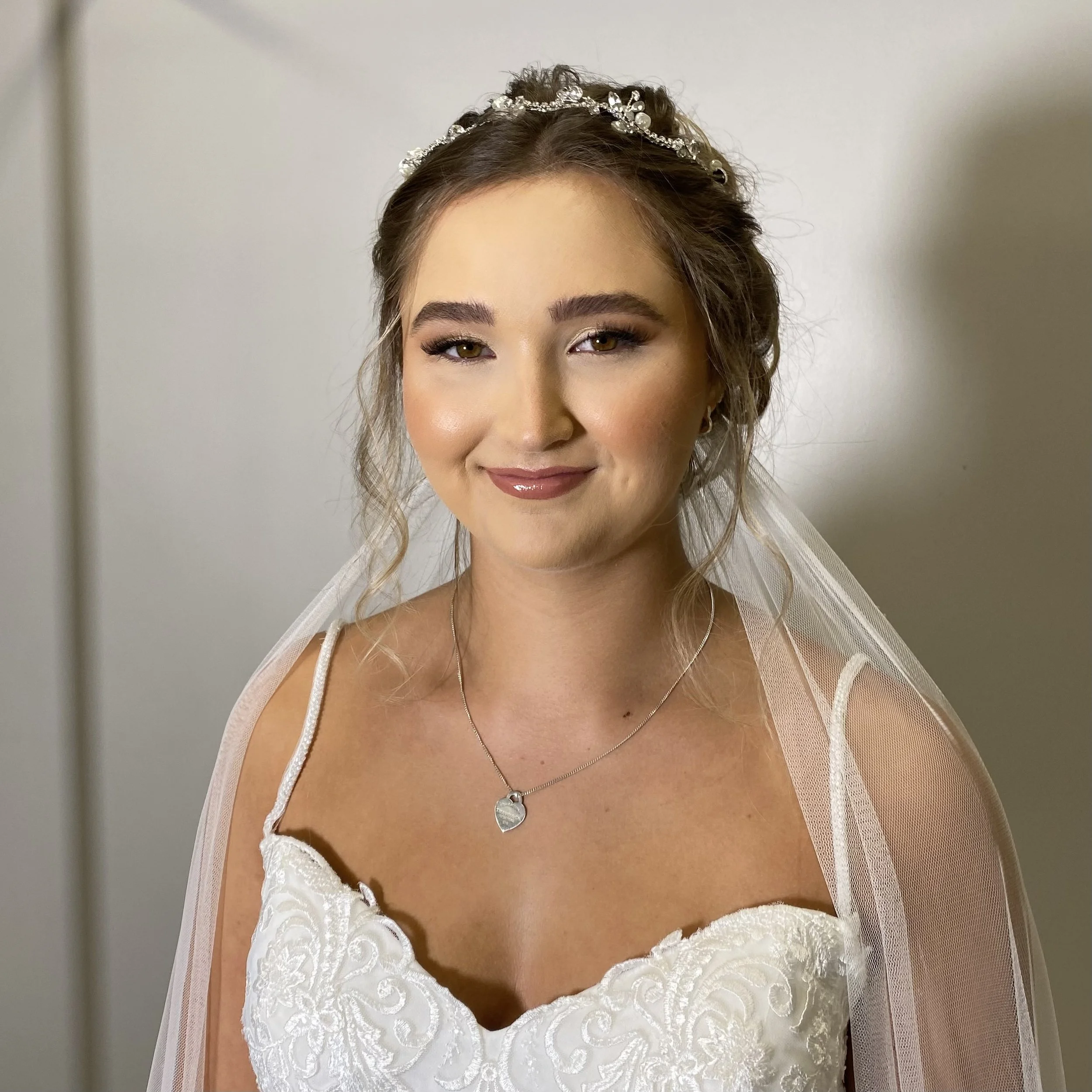 Close-up of a smiling woman with makeup and a tiara, wearing a wedding dress and jewelry, standing against a plain background.