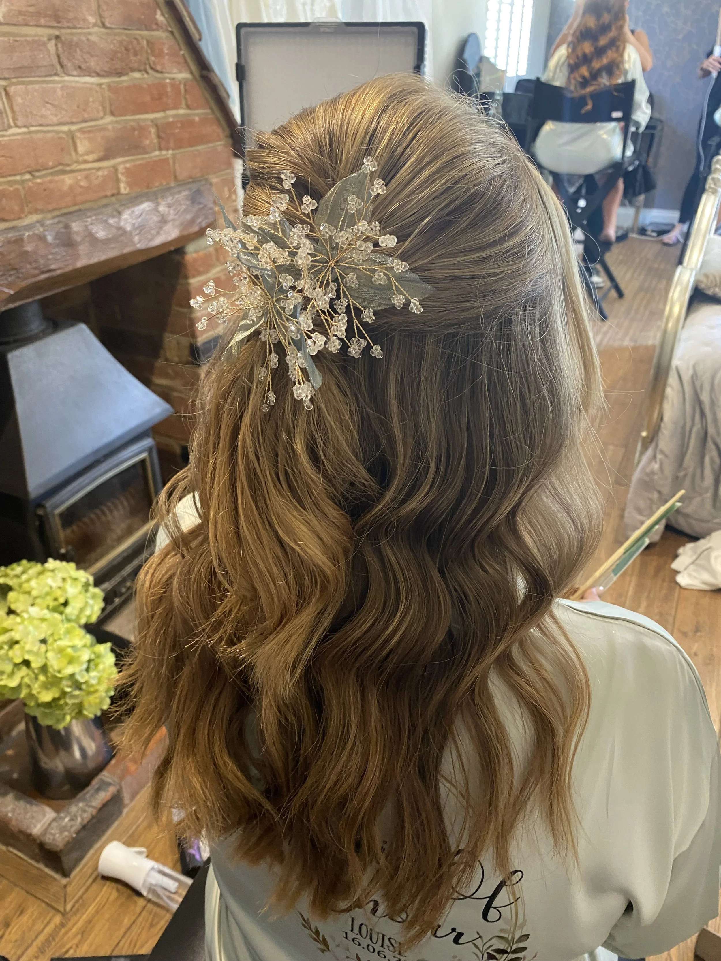 A woman with long, wavy, light brown hair styled with a decorative hairpiece featuring leaves and small clear beads, sitting indoors in front of a brick fireplace and a mirror.