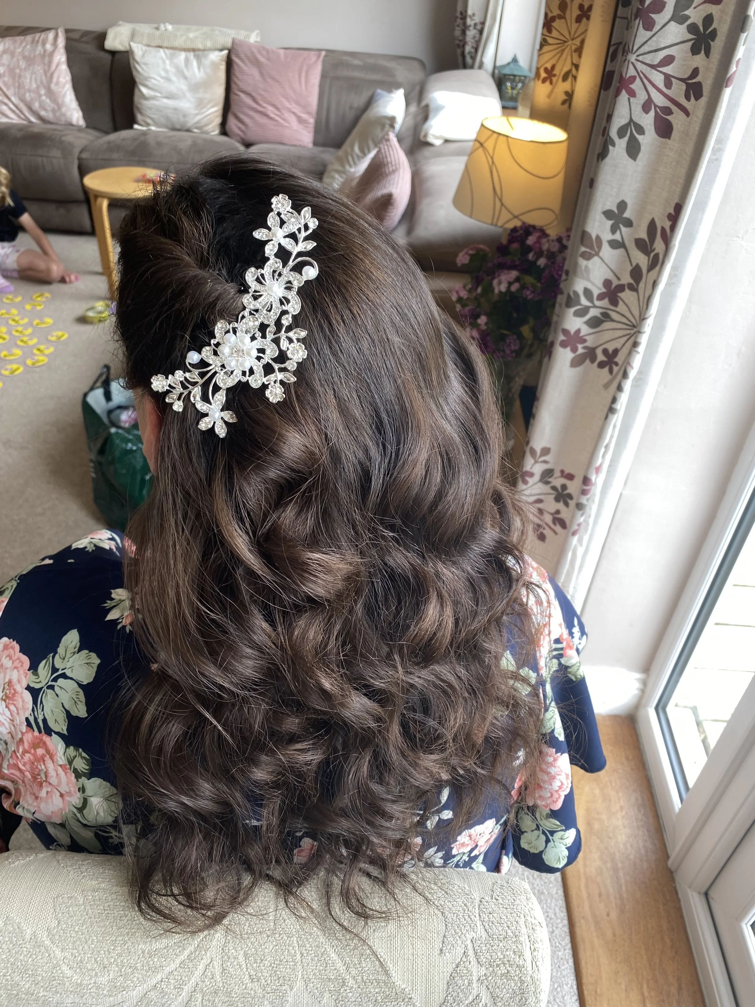 A woman with long, brown, curly hair wearing a decorative silver and pearl hair accessory, sitting on a light-colored, textured armchair in a living room.