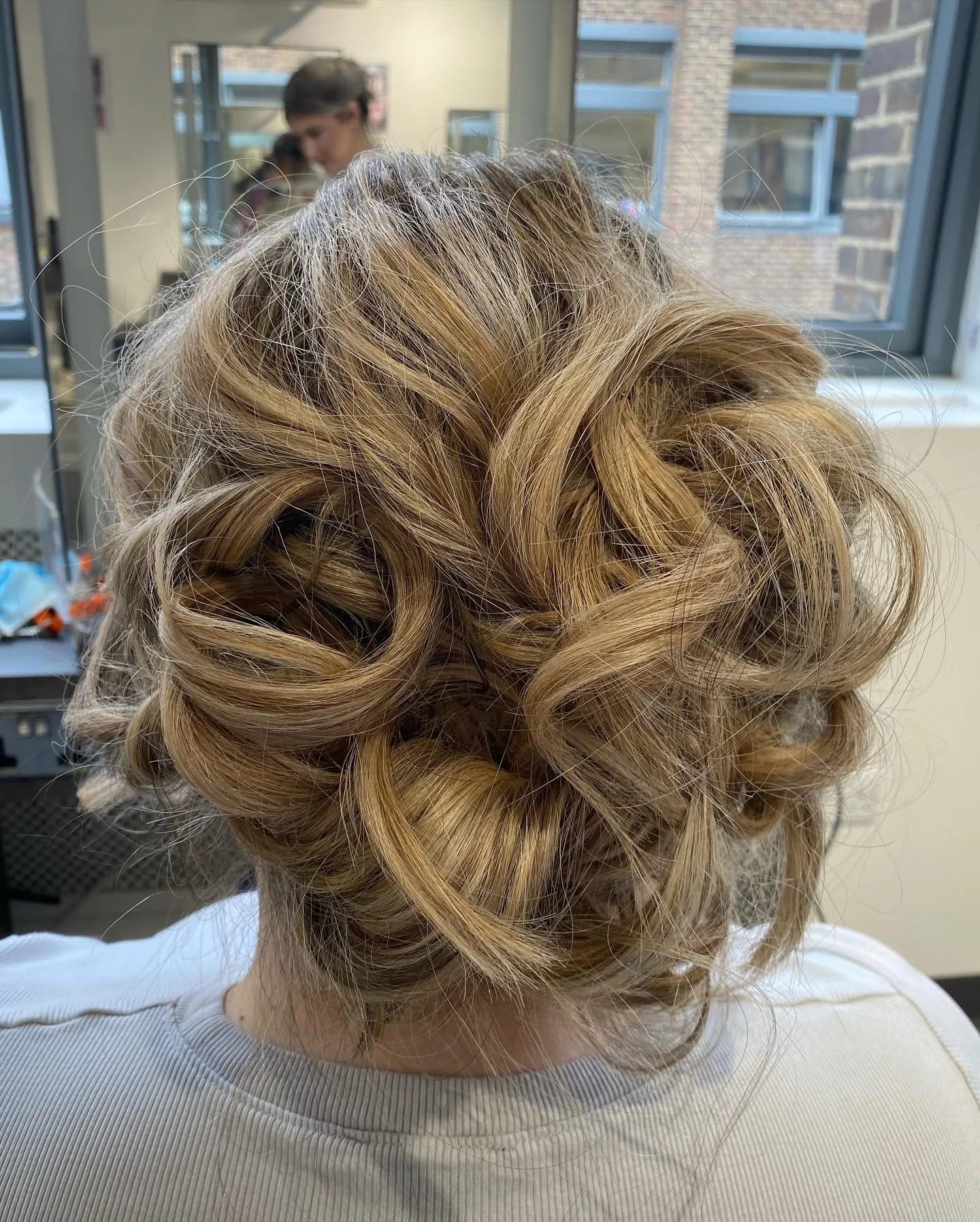 Close-up of a woman with styled blonde hair in an updo with tight curls. The background shows a window and some people in a room.