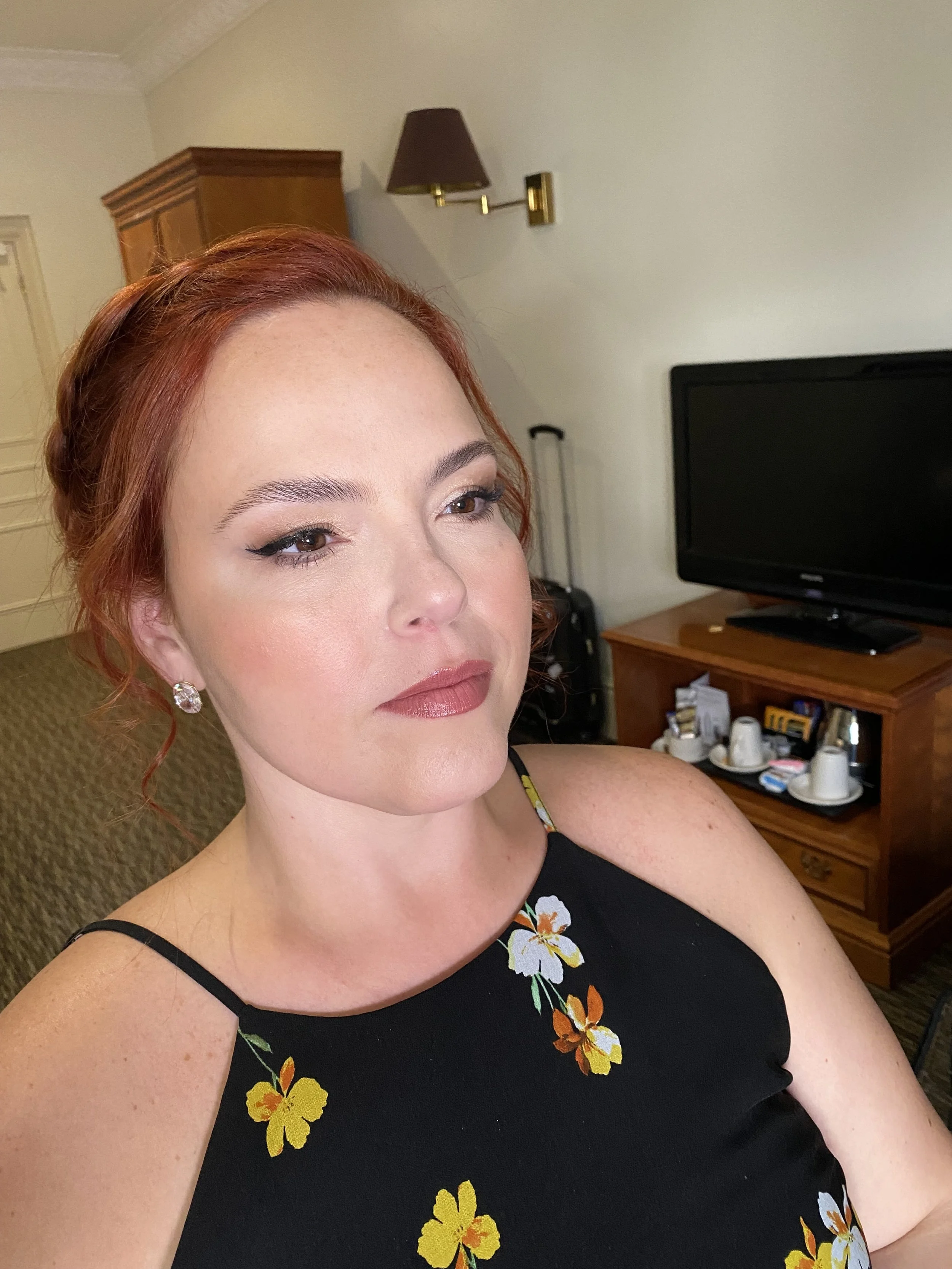 A woman with red hair and makeup, wearing a black dress with yellow and white floral patterns, taking a selfie in a hotel room.